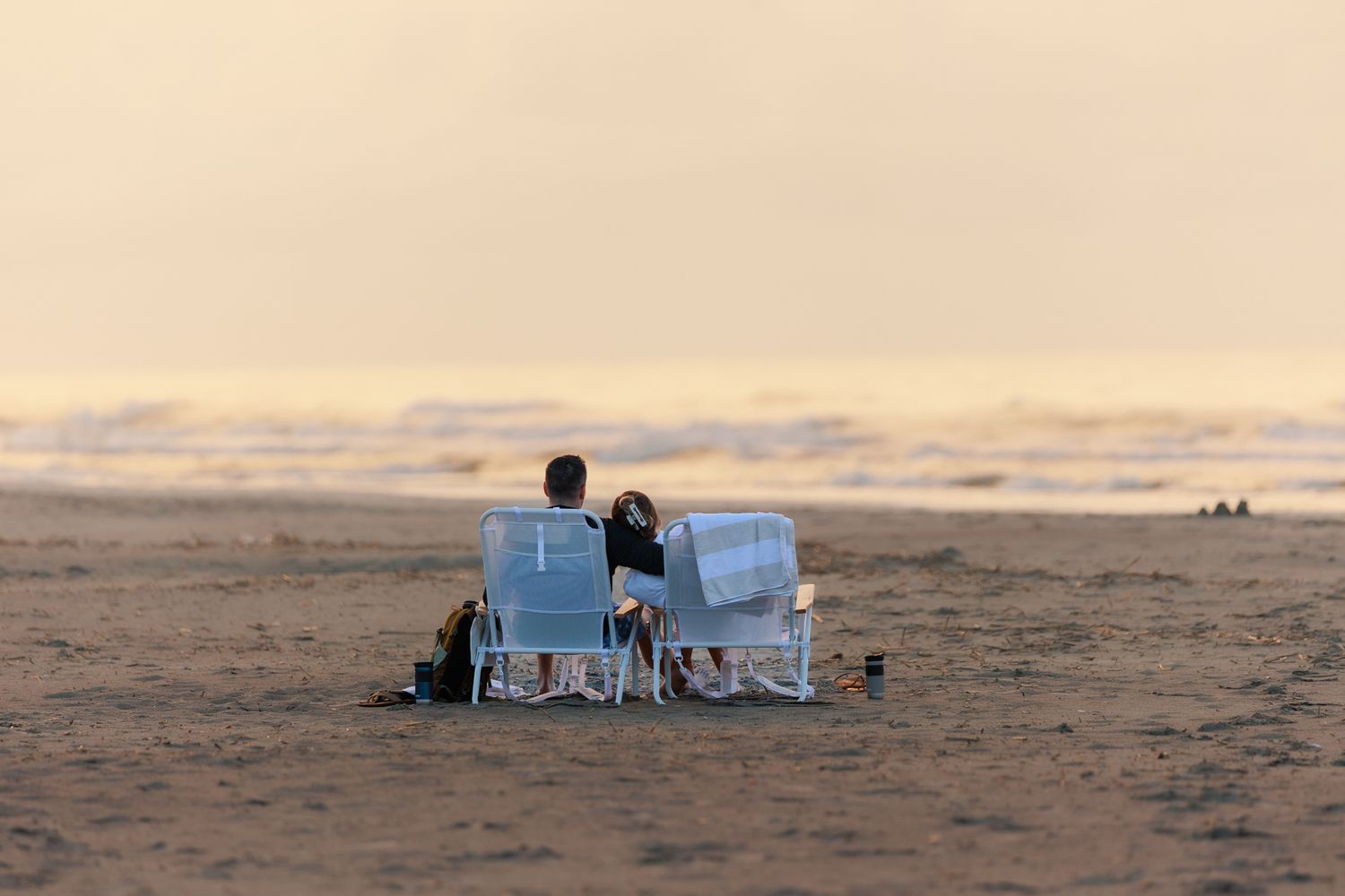 Couple sitting in beach chairs facing the ocean waves at sunset on a sandy beach.