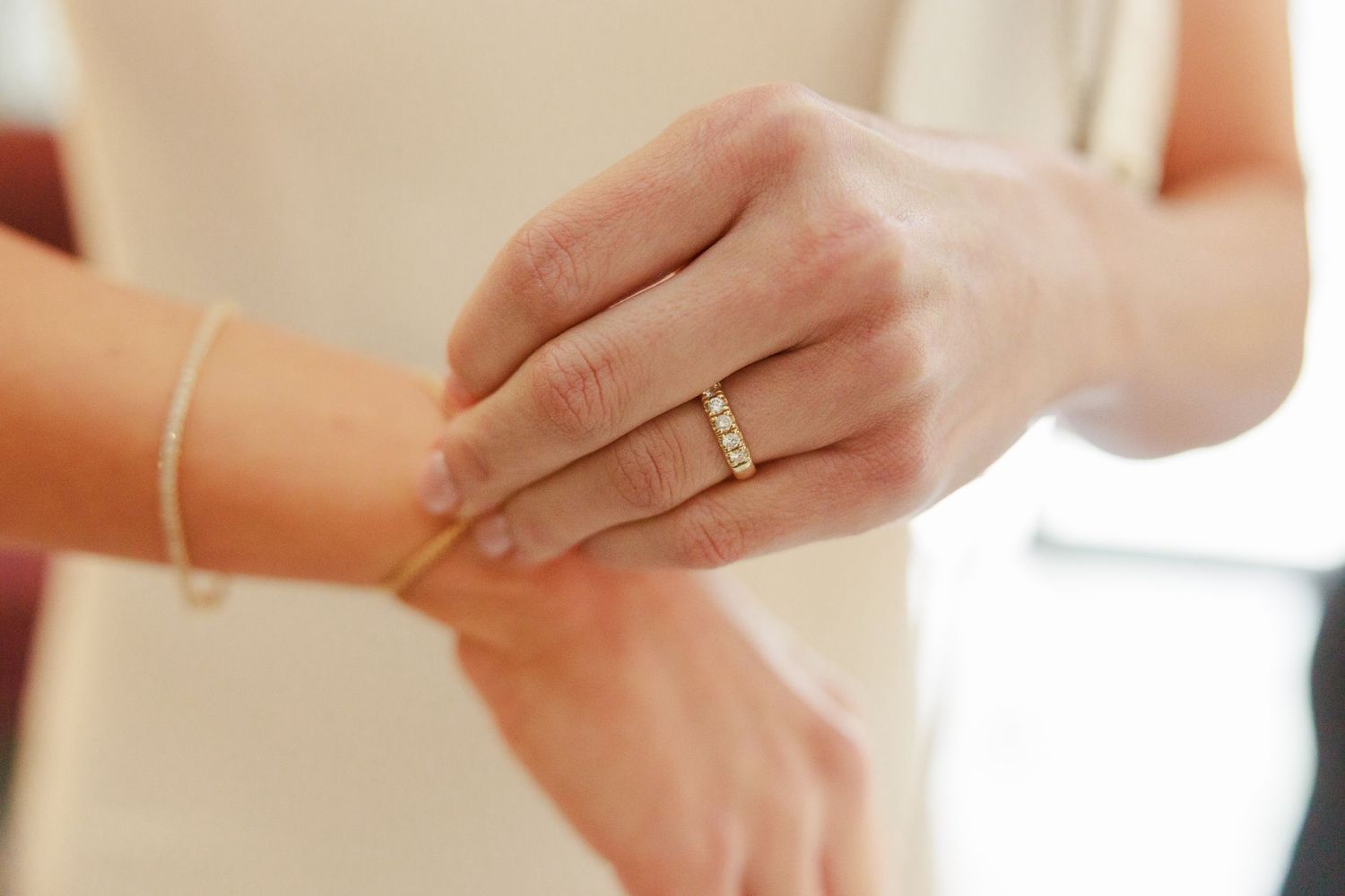 Close-up of a bride wearing a gold diamond eternity band ring, with another person fastening a gold bracelet.