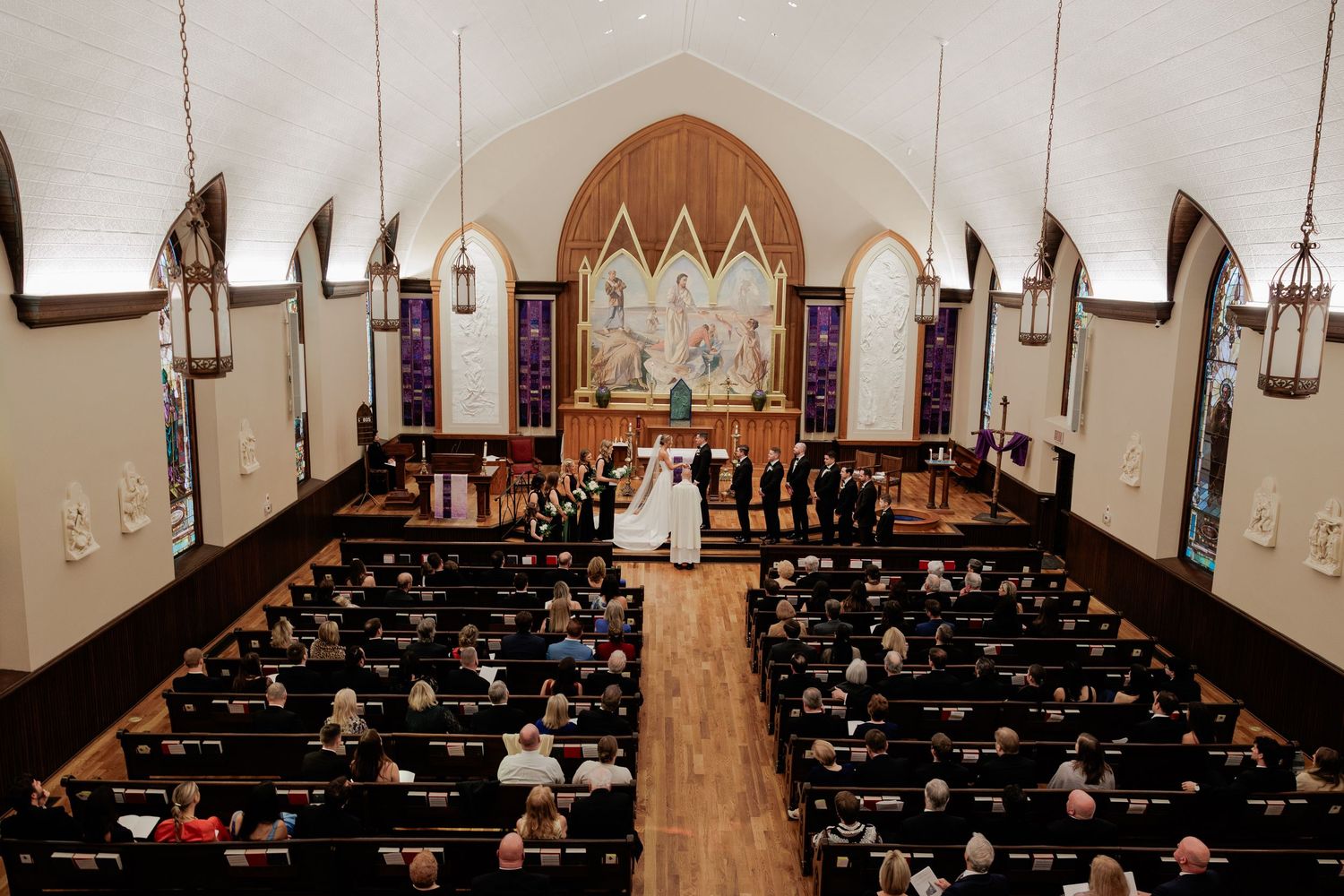 Elegant church wedding ceremony with guests seated in wooden pews, couple at altar beneath stained glass windows.