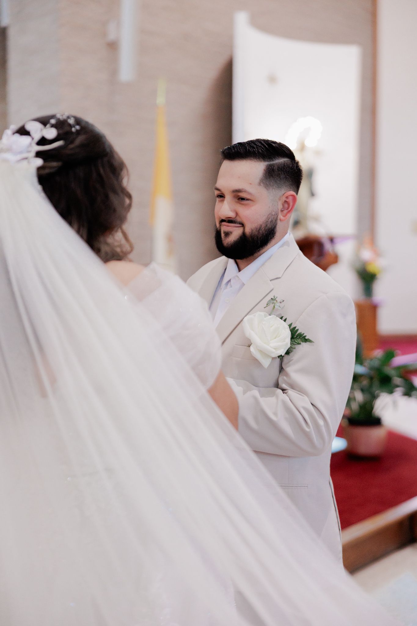 Groom in beige suit with white boutonniere smiling at bride in veil during church wedding ceremony.
