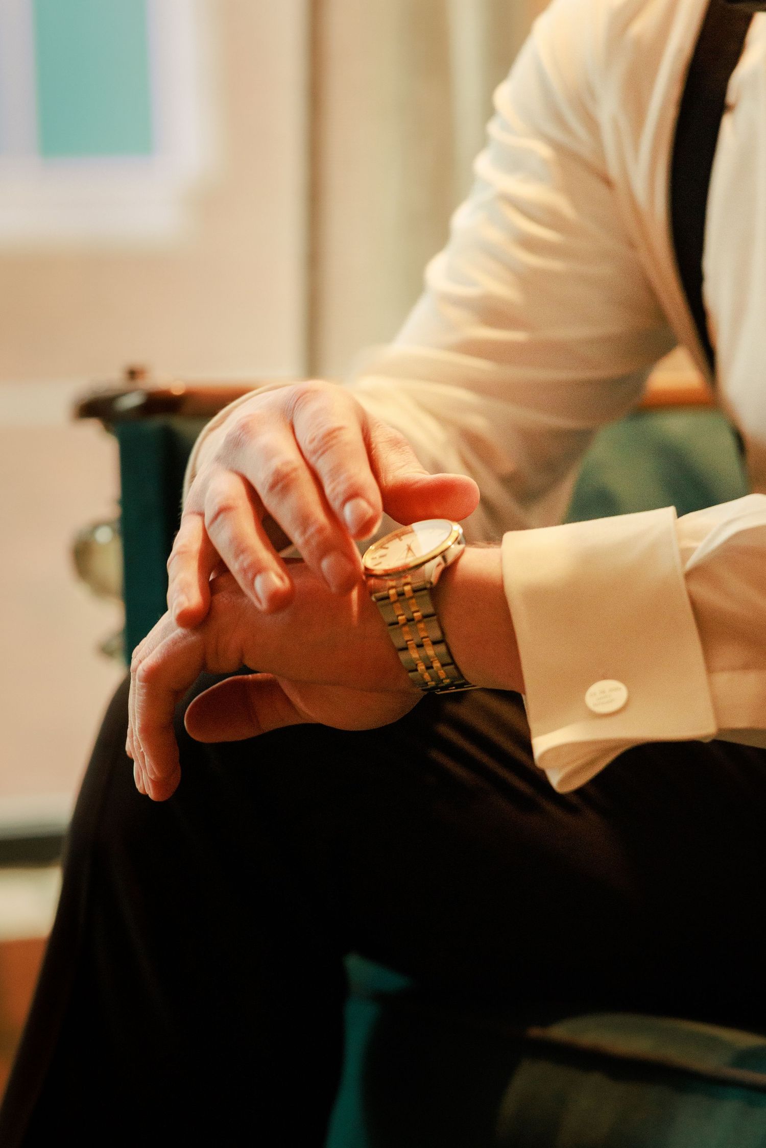 Man in white dress shirt with gold watch and suspenders sitting with hands clasped, elegant formal attire.