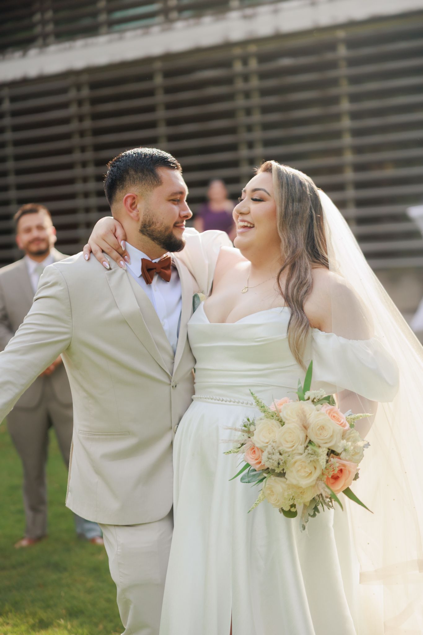Smiling bride in white gown with bouquet and groom in beige suit with burgundy bow tie share a joyful moment outdoors.