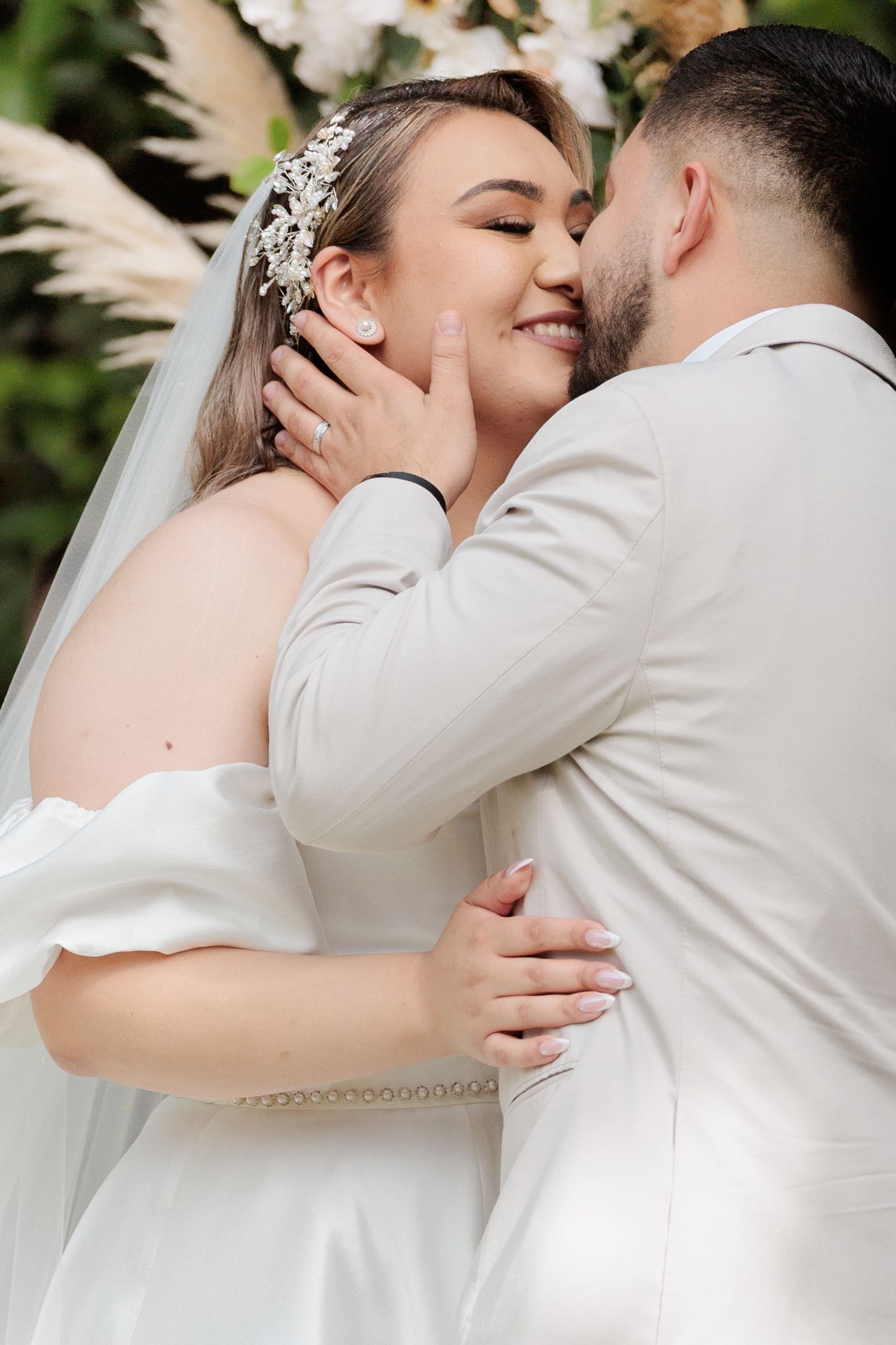 A bride and groom share a romantic kiss at their wedding ceremony, with the bride wearing a white gown and floral hair accessory.