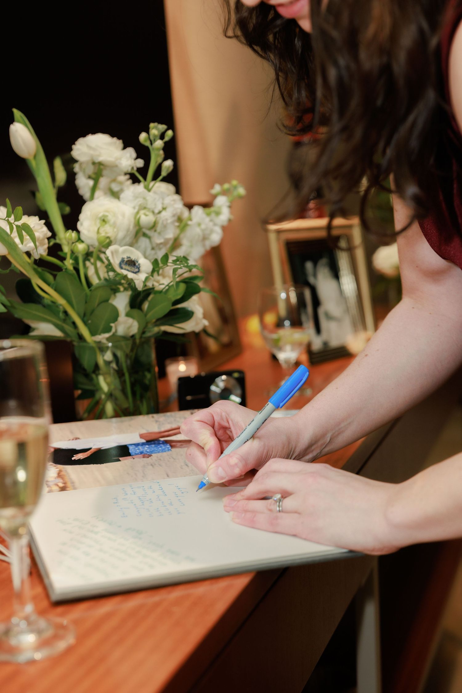 Woman in red dress signing a wedding guest book with a blue pen beside white flowers and champagne glasses.