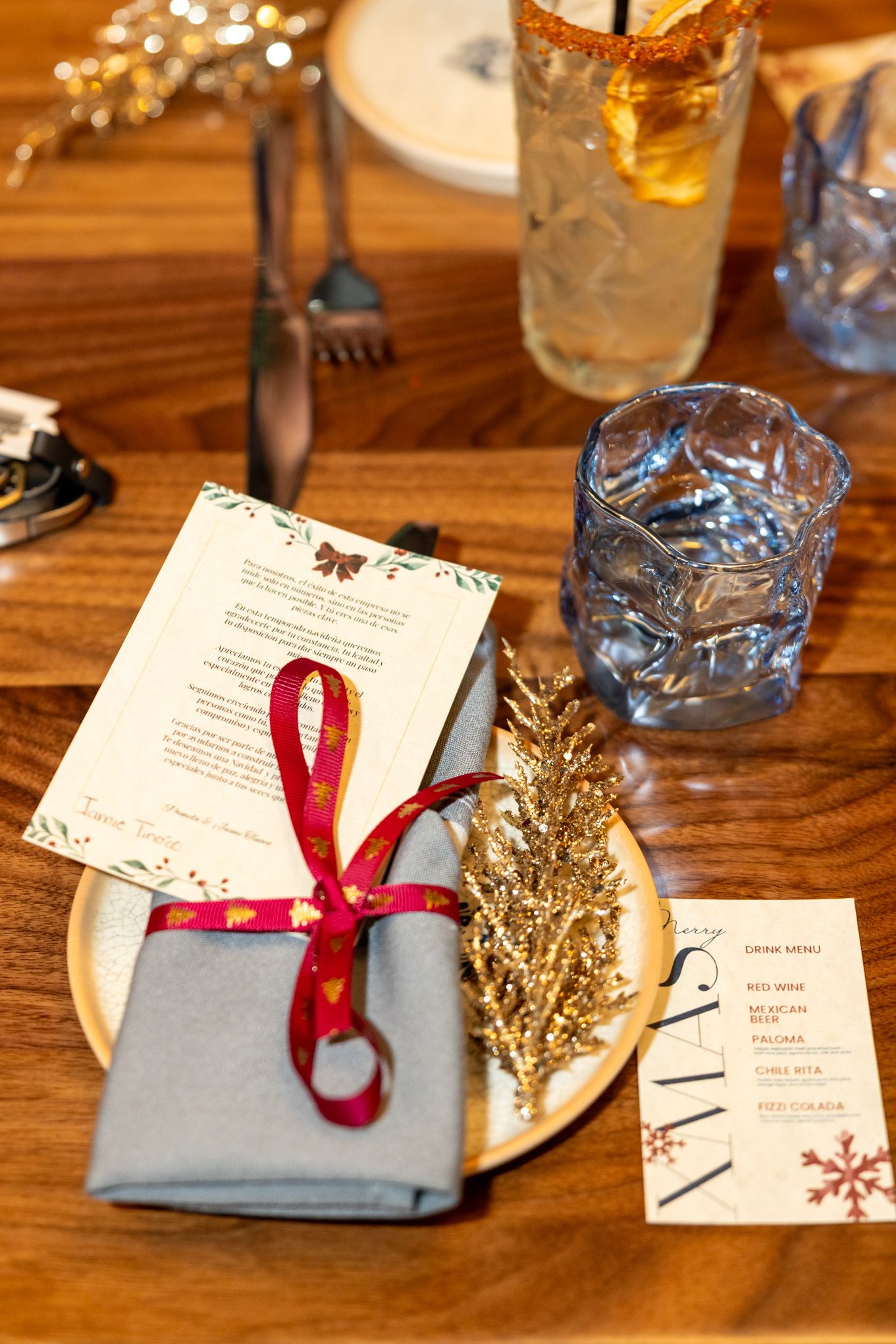 Festive Christmas table setting with gold tree, red ribbon napkin, holiday menu card, and blue glassware on wooden table.