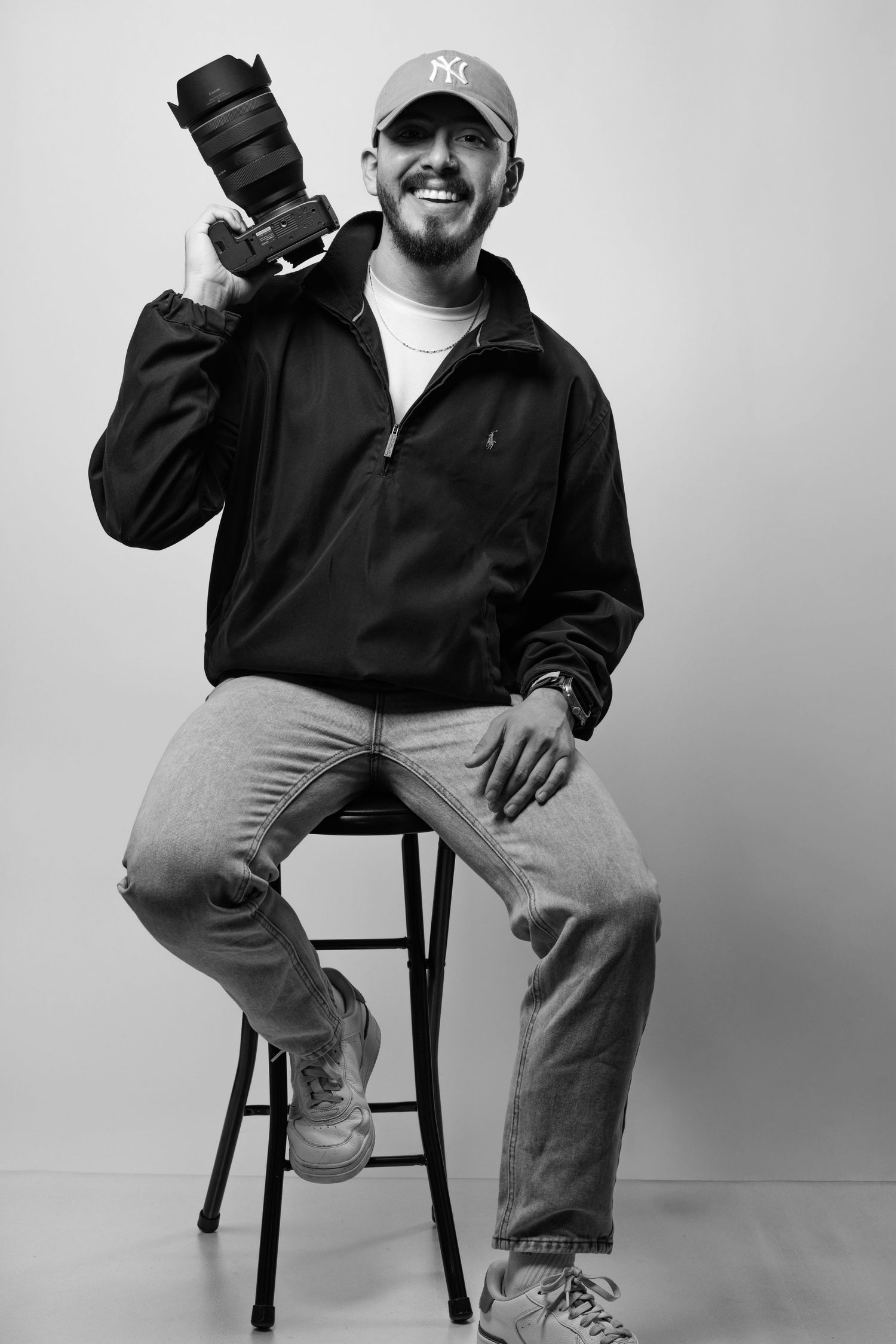 Black and white portrait of a photographer sitting on a stool holding a professional camera and wearing a NY Yankees cap.