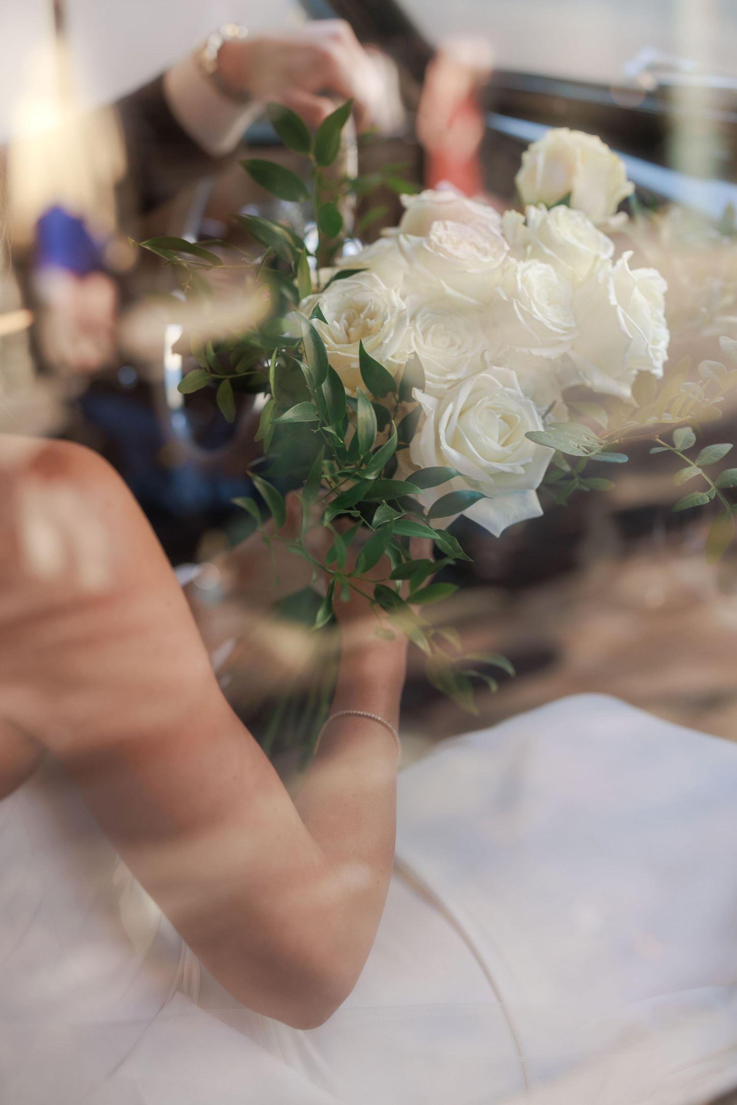 Bride holding a elegant white rose bouquet with green leaves, captured through glass reflection at a wedding ceremony.