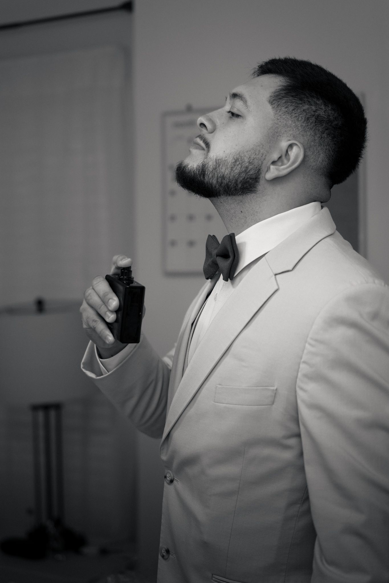 Black and white photo of a groom in a suit and bow tie applying cologne before his wedding ceremony.