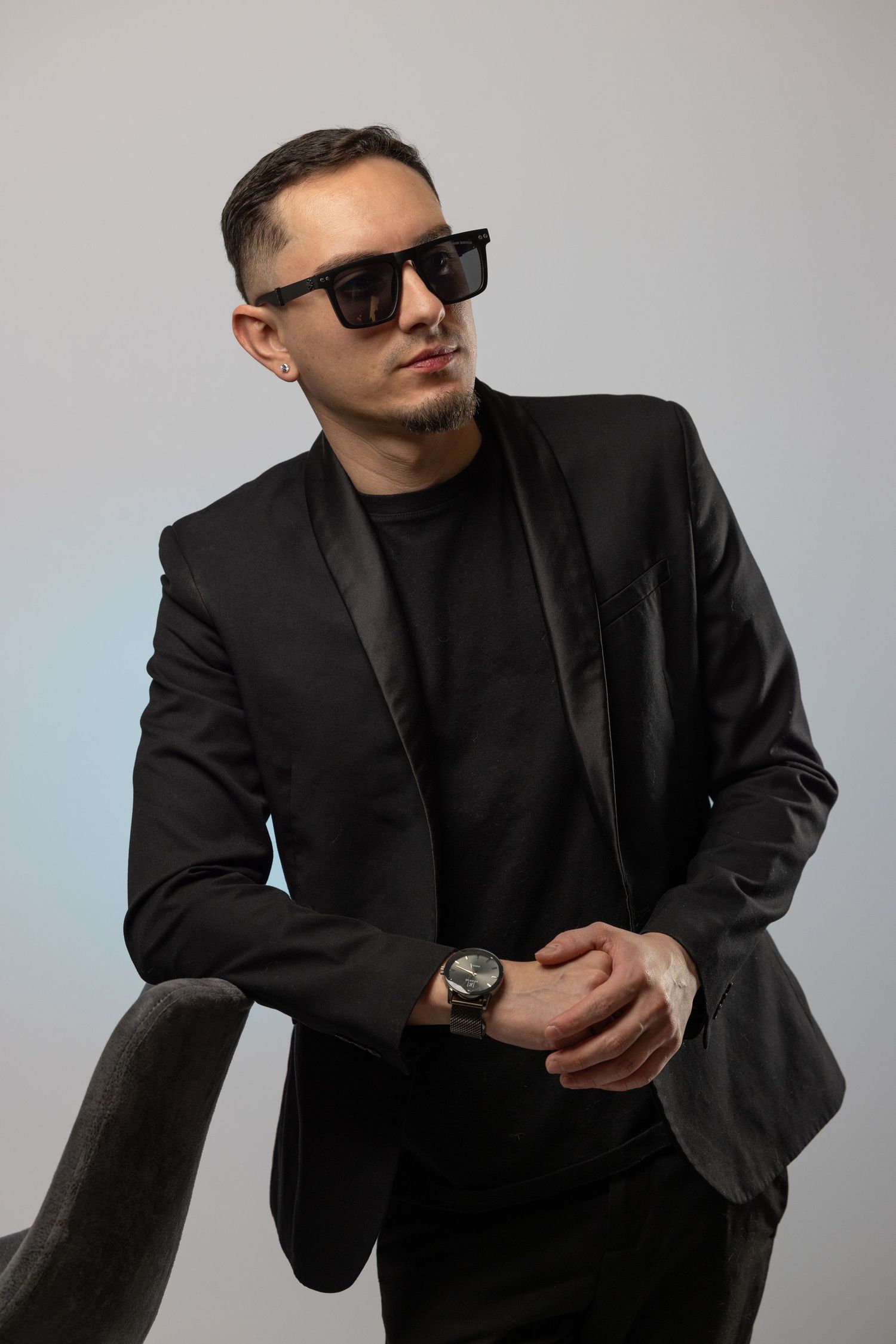 Stylish man in black suit and sunglasses wearing a watch, posing against a light gray background.