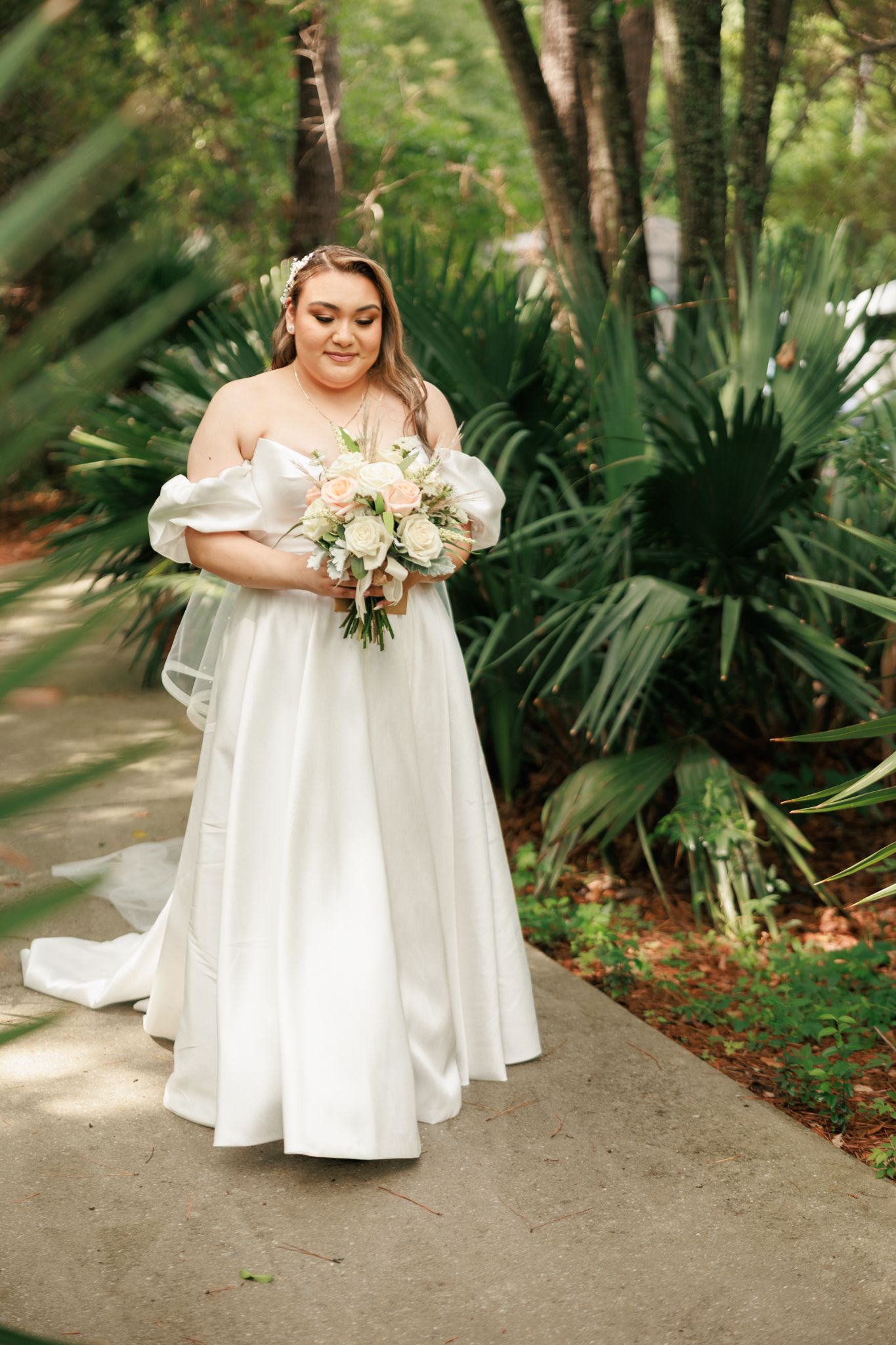 Bride in off-shoulder white gown holding floral bouquet among lush tropical greenery.