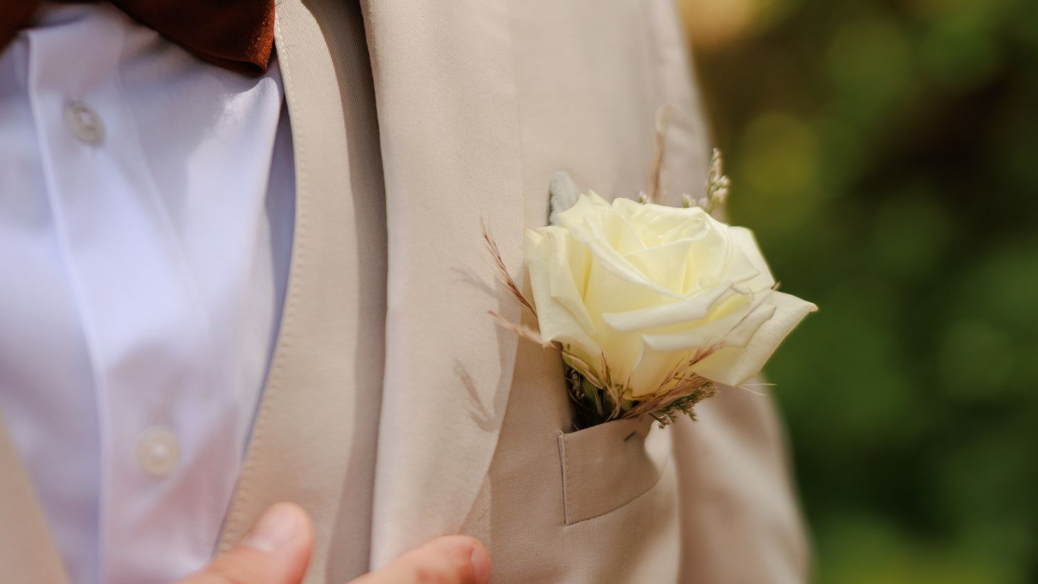 A groom wearing a beige suit with a white rose boutonniere pinned to his lapel at an outdoor wedding ceremony.