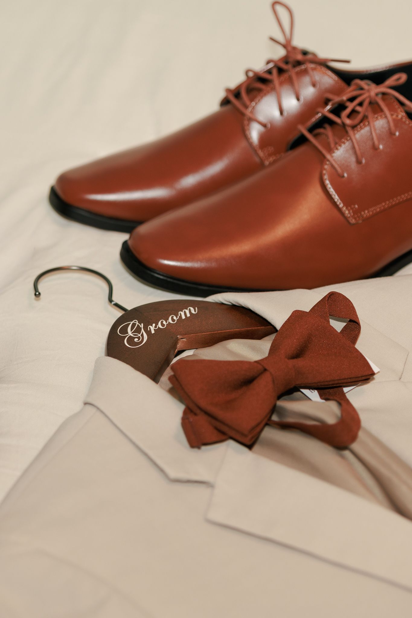 Groom wedding accessories flat lay with brown leather dress shoes, rust bow tie, and white shirt on a hanger.
