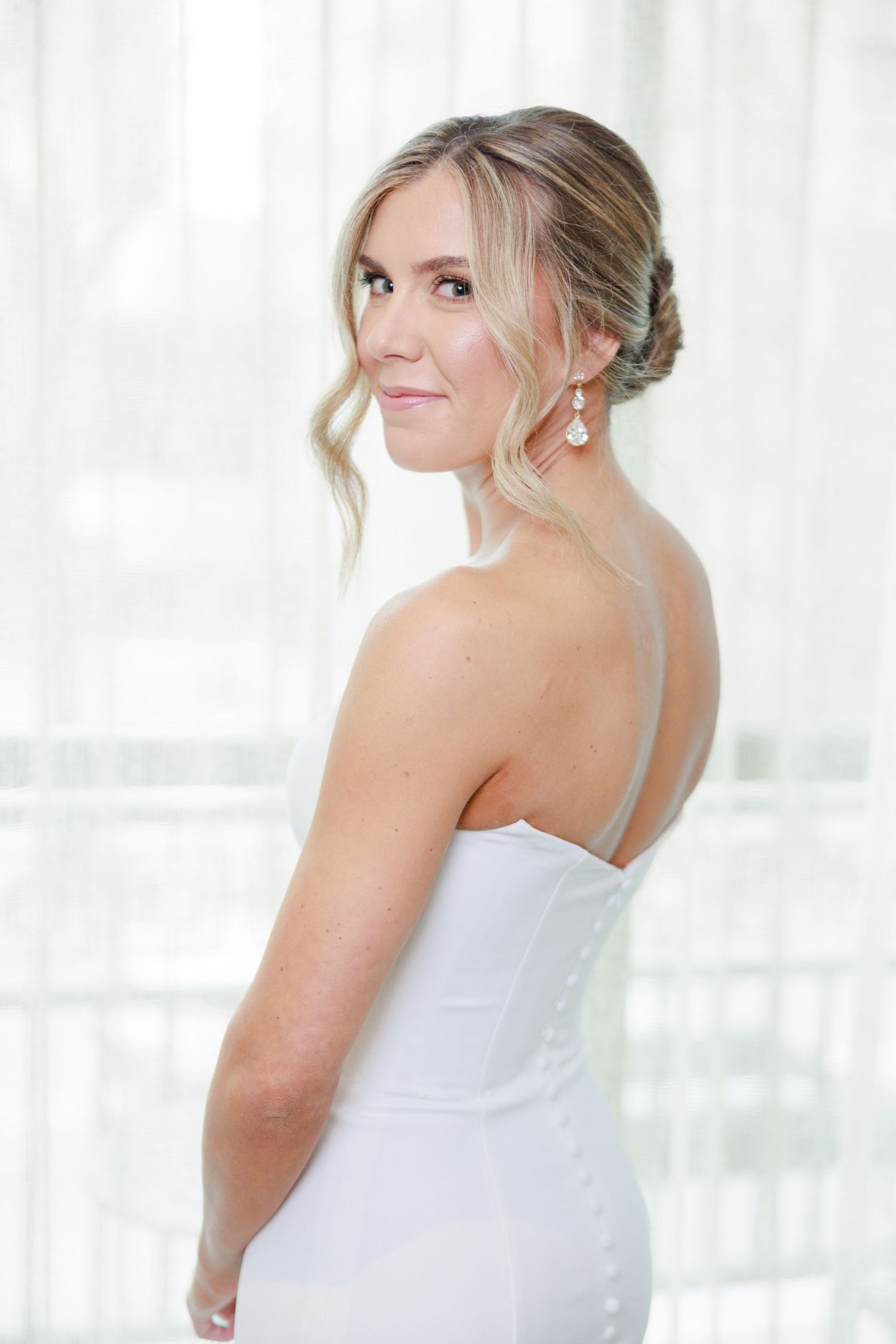 Blonde bride in a white strapless gown with elegant updo and drop earrings, posing gracefully in soft natural light.