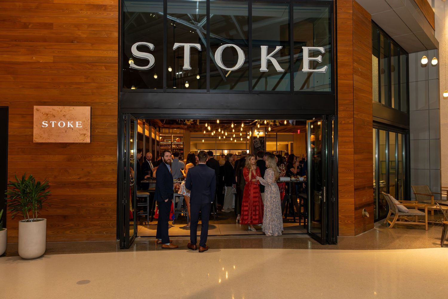 Guests gather at the entrance of Stoke restaurant, featuring warm wood accents and string lights inside.