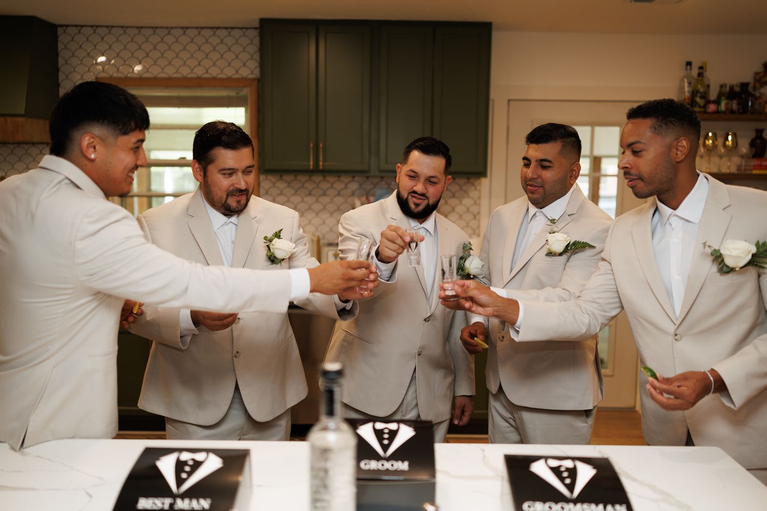 Five groomsmen in beige suits toasting drinks at a wedding, with Best Man and Groomsman signs on the counter.