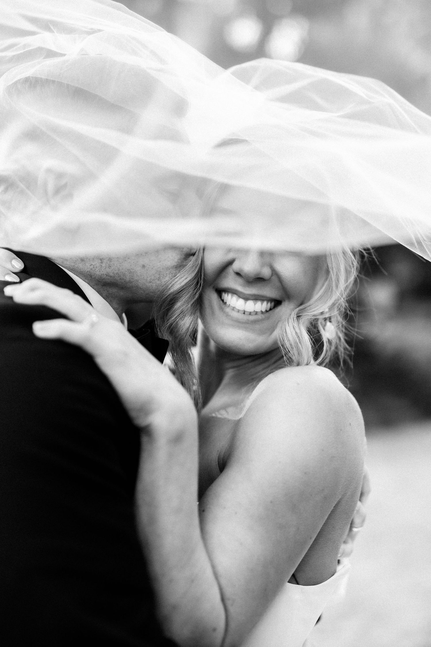 Black and white photo of a smiling bride under her veil being kissed by groom on wedding day.