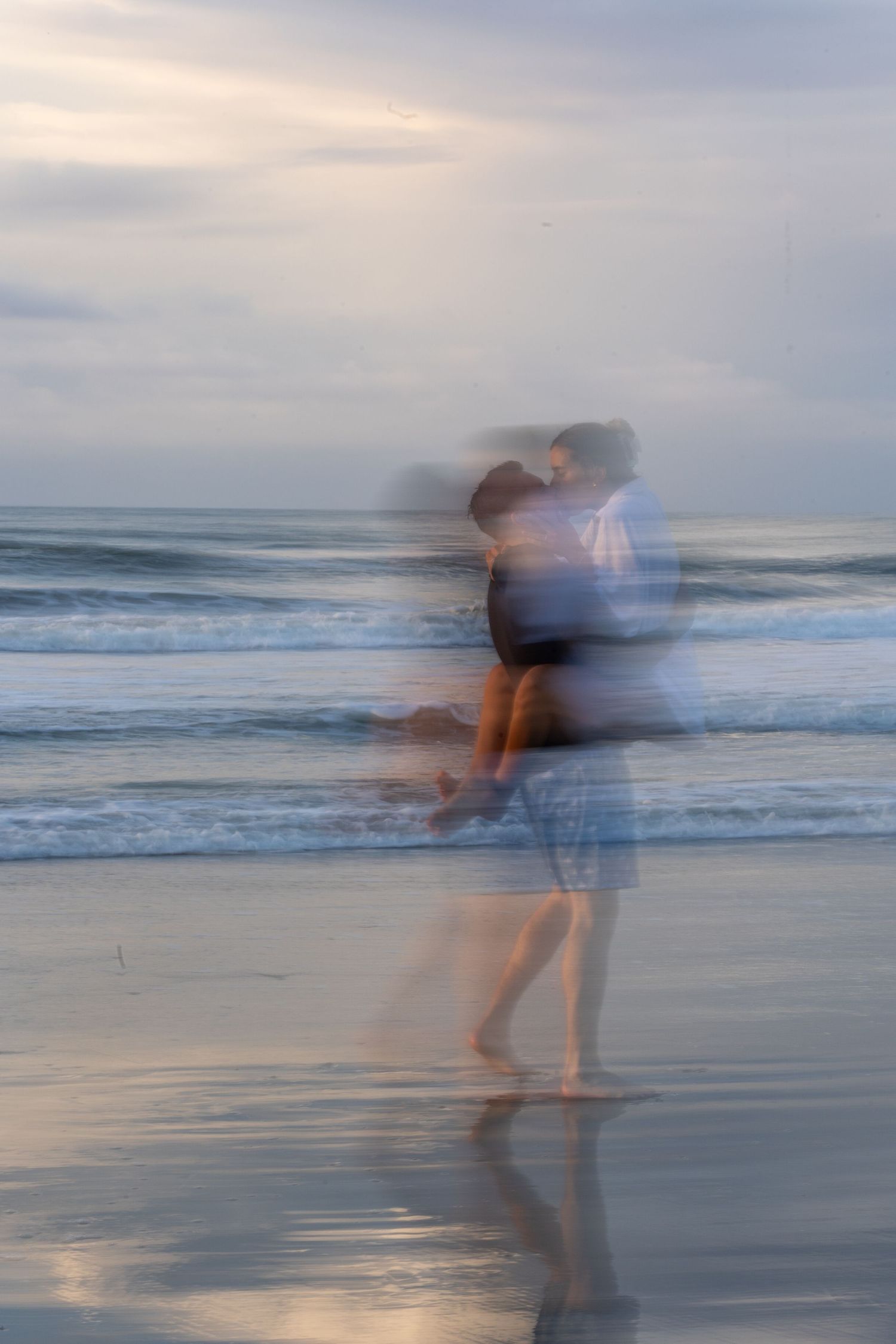 Motion blur photograph of a couple dancing on a beach at sunset with soft waves in the background.
