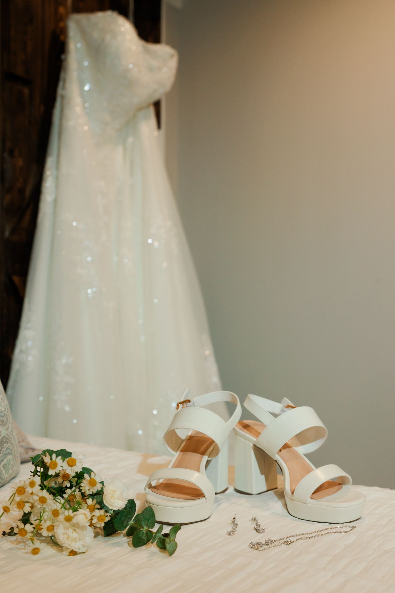 White bridal platform heels, floral bouquet, and earrings displayed on a table with a sparkling wedding gown hanging in the background.