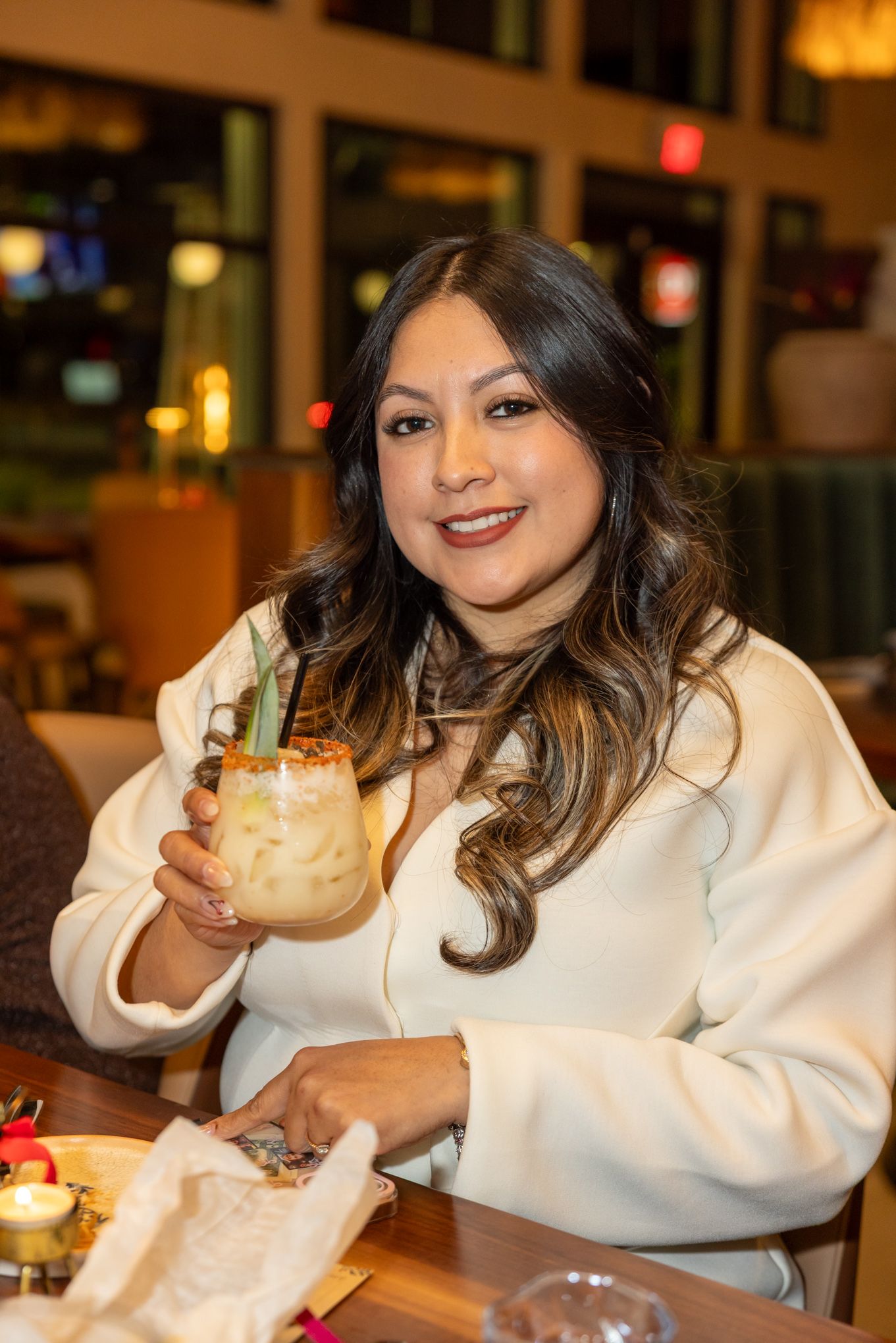 Smiling woman in white jacket holding a tropical cocktail with pineapple garnish at a restaurant.