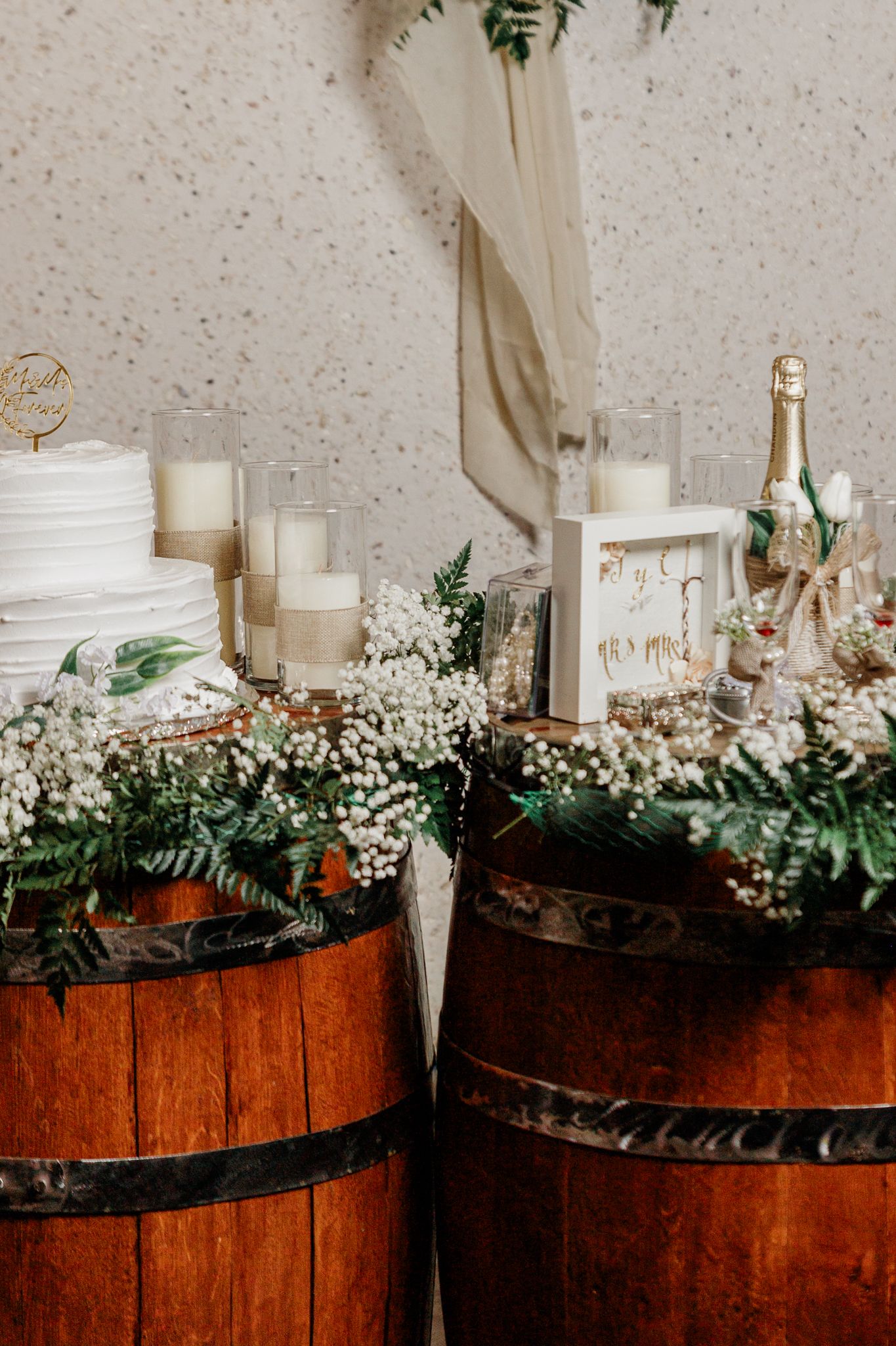 Rustic wedding cake display on wooden barrels with white florals, candles, champagne, and greenery decor.