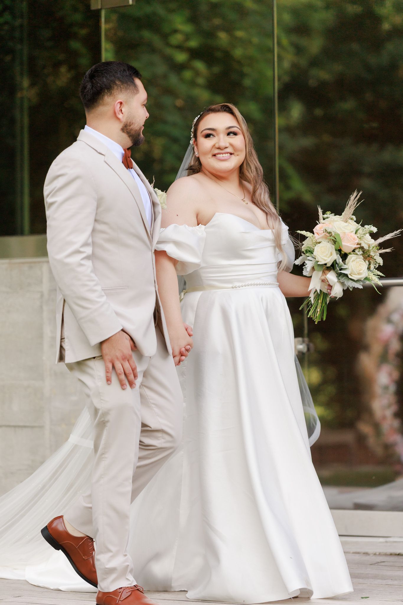 Smiling bride in white off-shoulder gown holds bouquet while walking hand-in-hand with groom in beige suit outdoors.