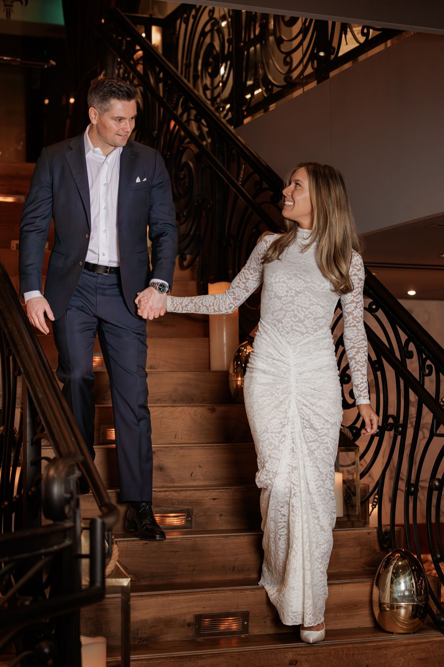 Couple in formal attire holding hands while descending an elegant staircase with ornate iron railings.