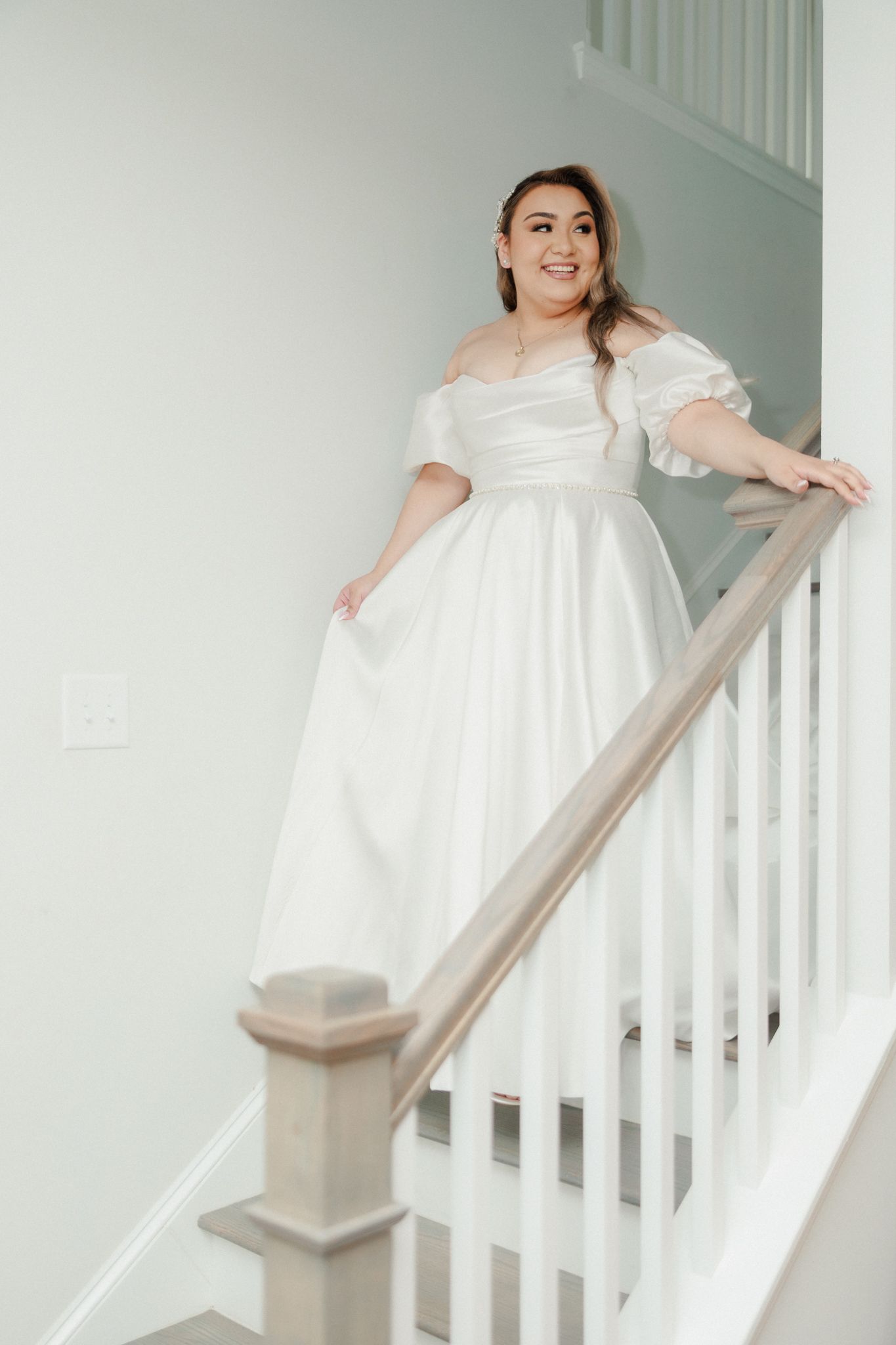 Smiling bride in off-shoulder white ball gown poses gracefully on staircase railing in bright home interior.