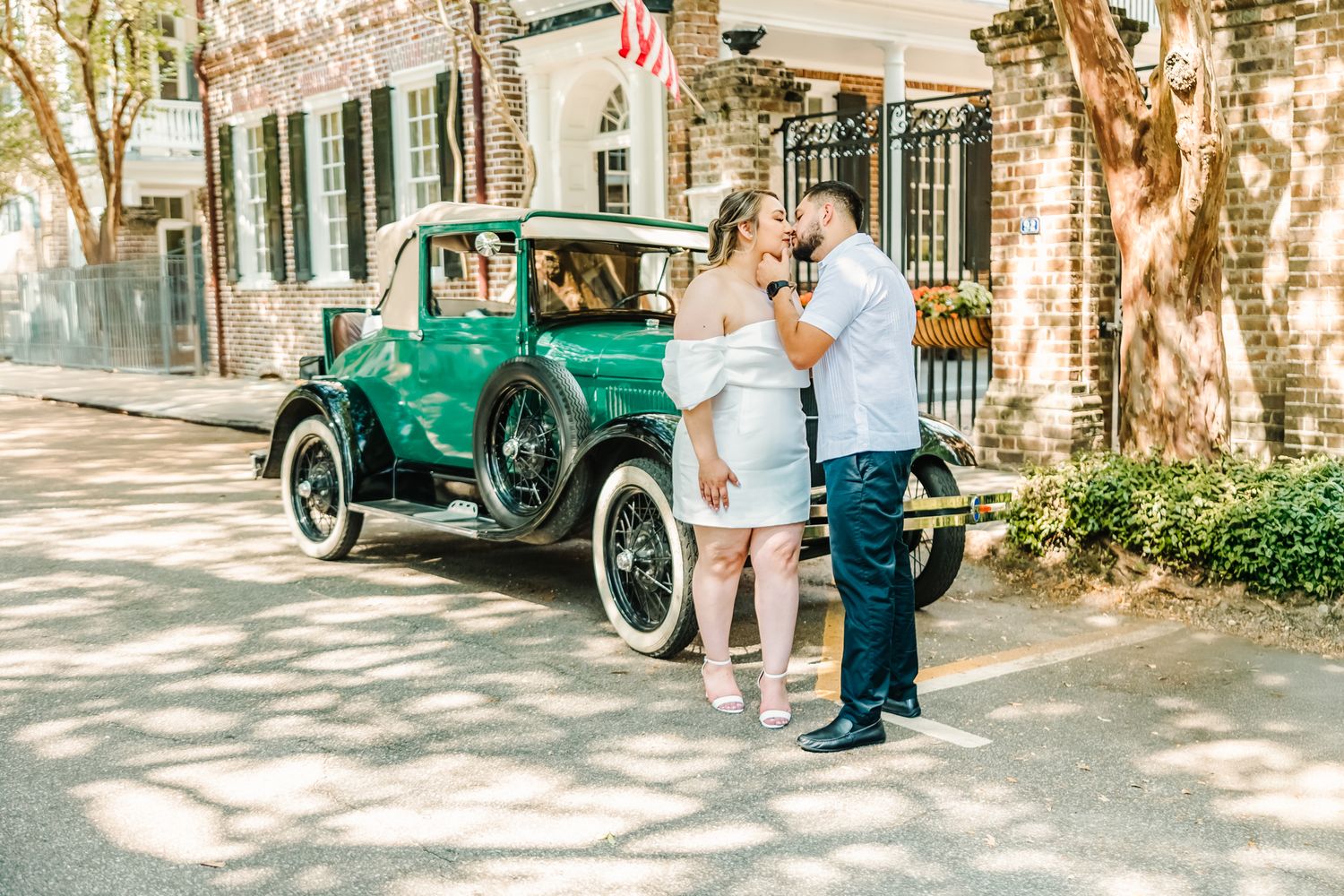 Couple sharing a romantic kiss beside a vintage green antique car on a sunny cobblestone street.