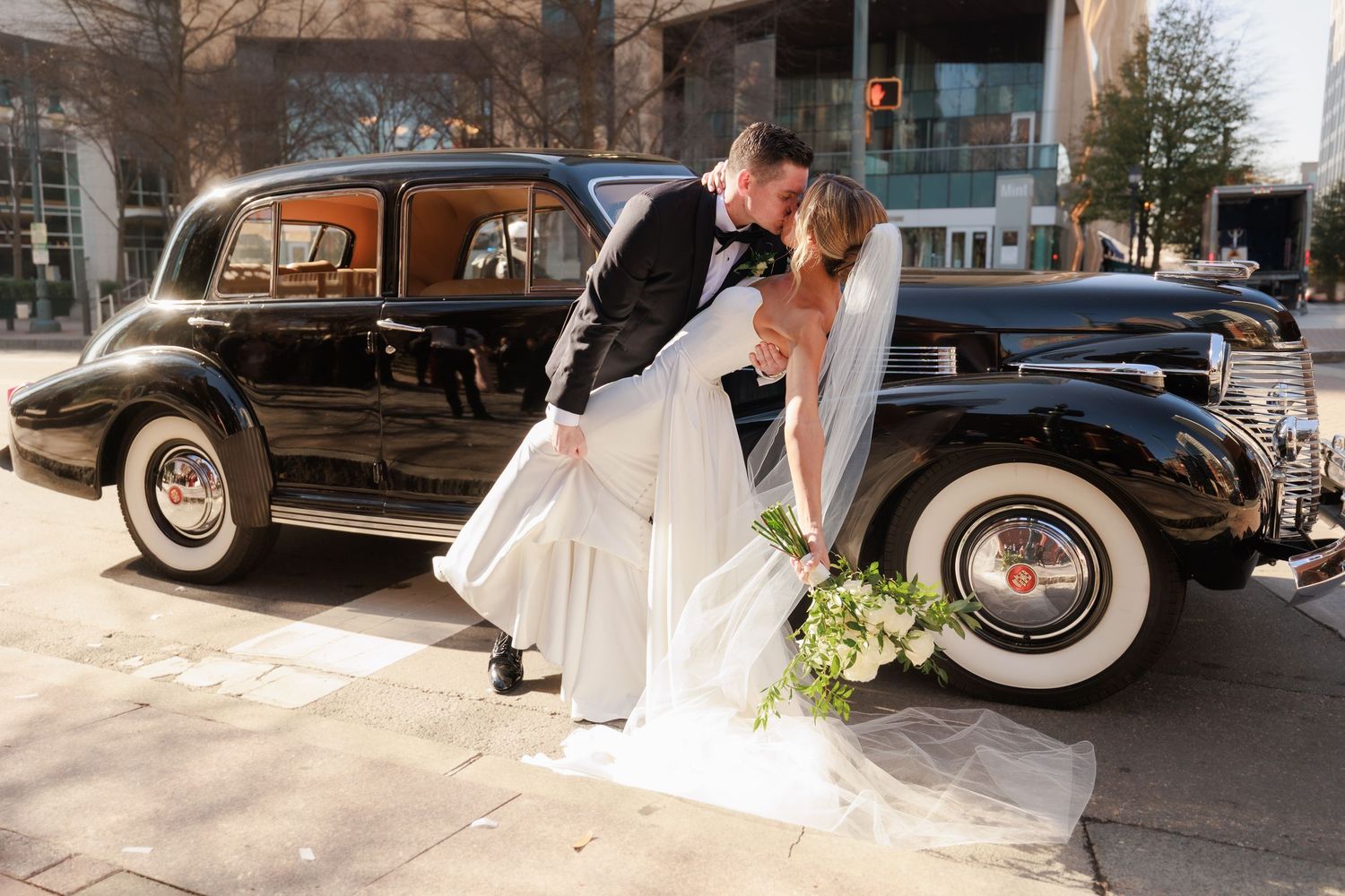 Bride and groom sharing a romantic dip kiss beside a classic black vintage car on their wedding day.