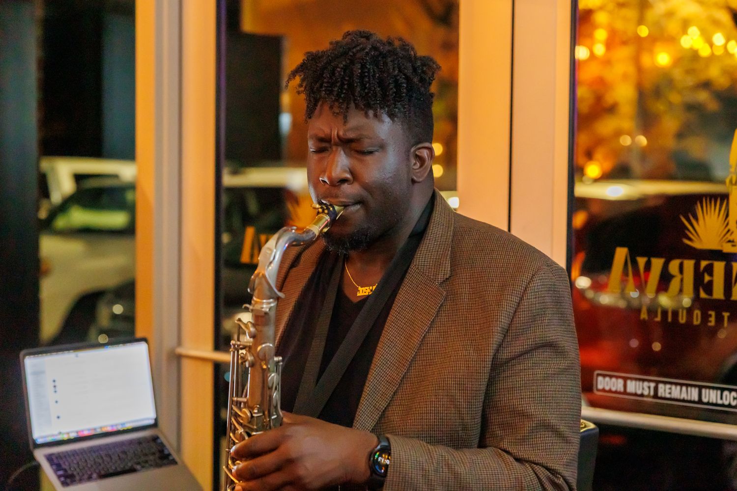 A musician plays saxophone outdoors at night, wearing a brown blazer, with a laptop visible nearby.