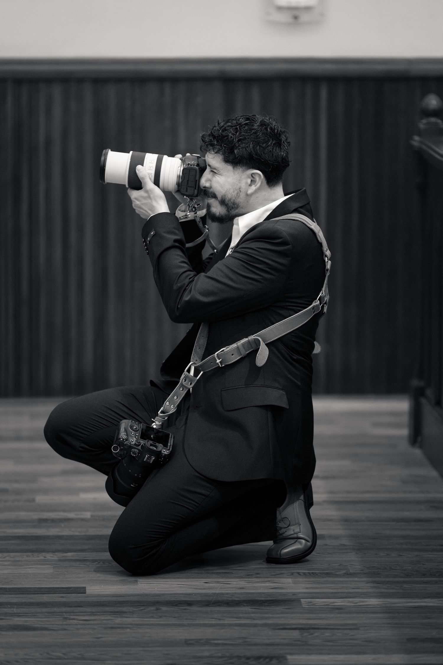 Black and white photo of a male photographer kneeling on floor, shooting with a large telephoto lens camera in formal attire.