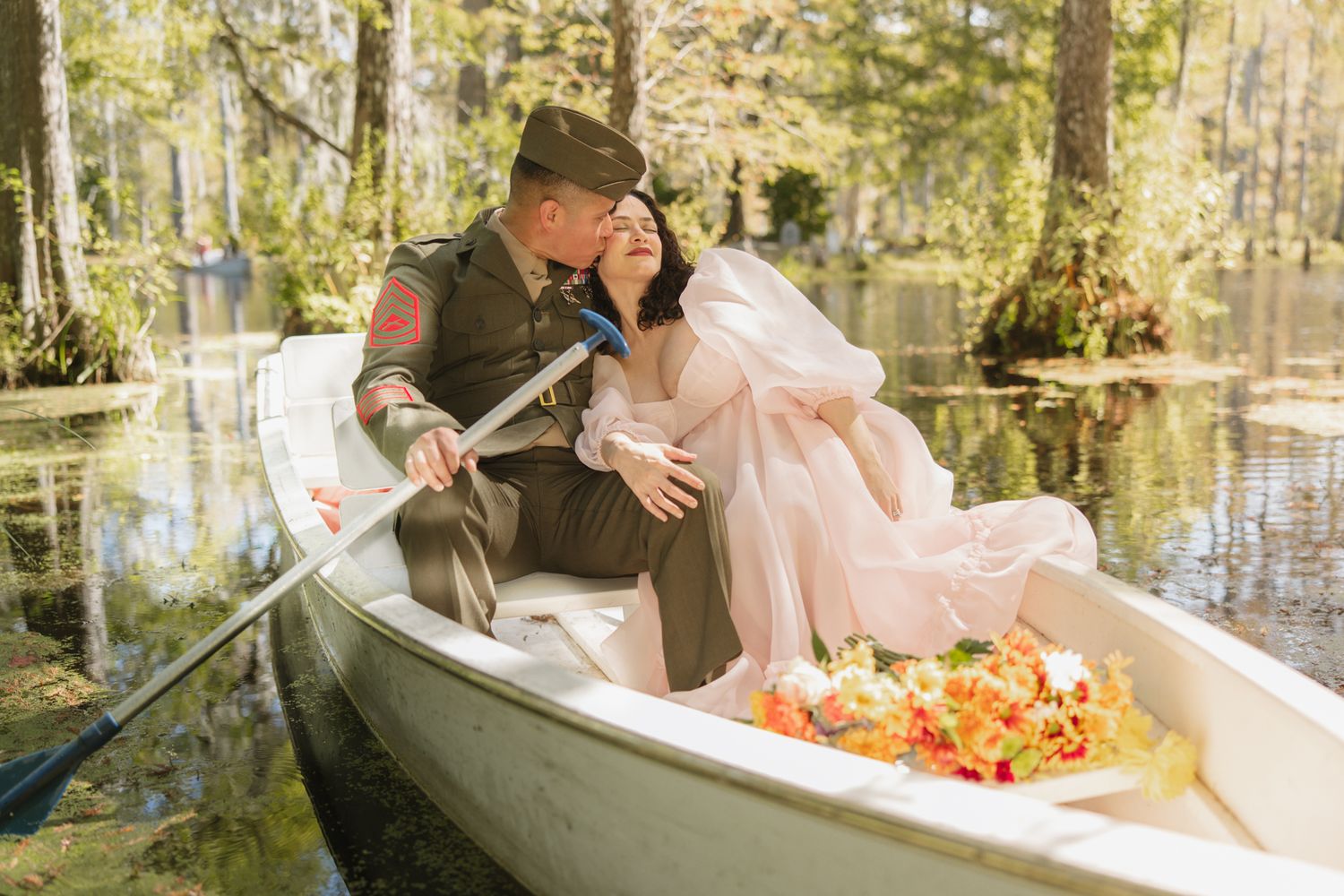 Military groom and bride in pink dress sharing romantic moment in rowboat on scenic cypress lake with floral bouquet.