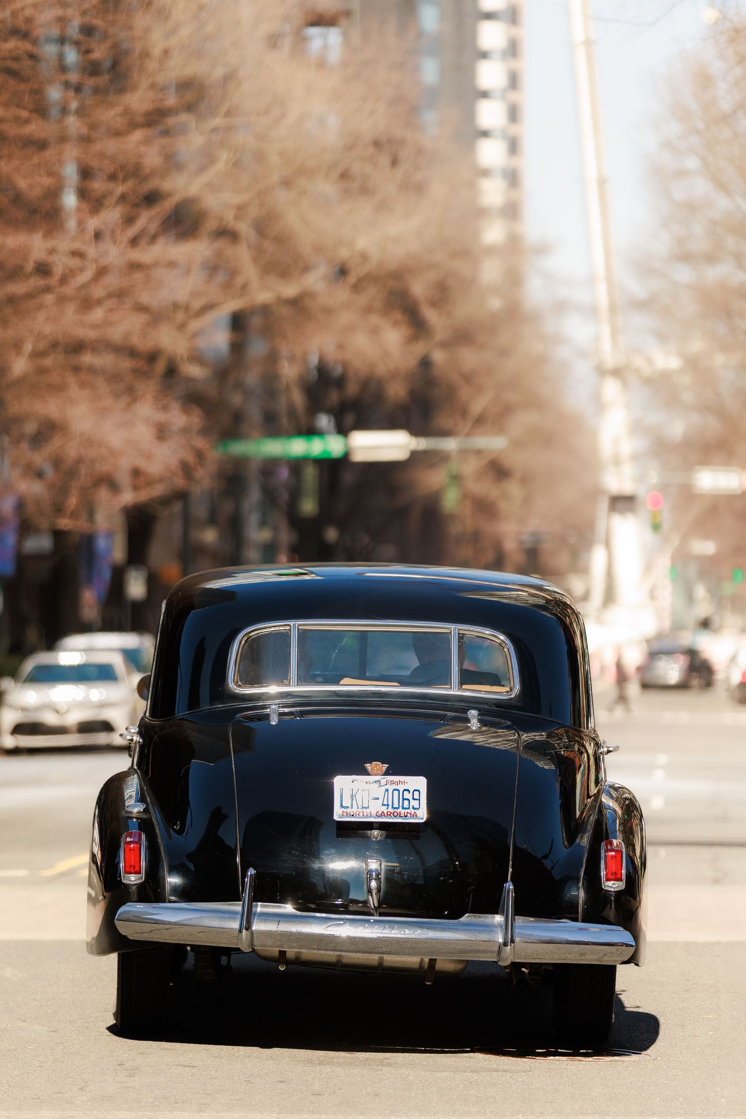 Rear view of a classic black vintage car driving down a city street with bare trees and traffic lights.