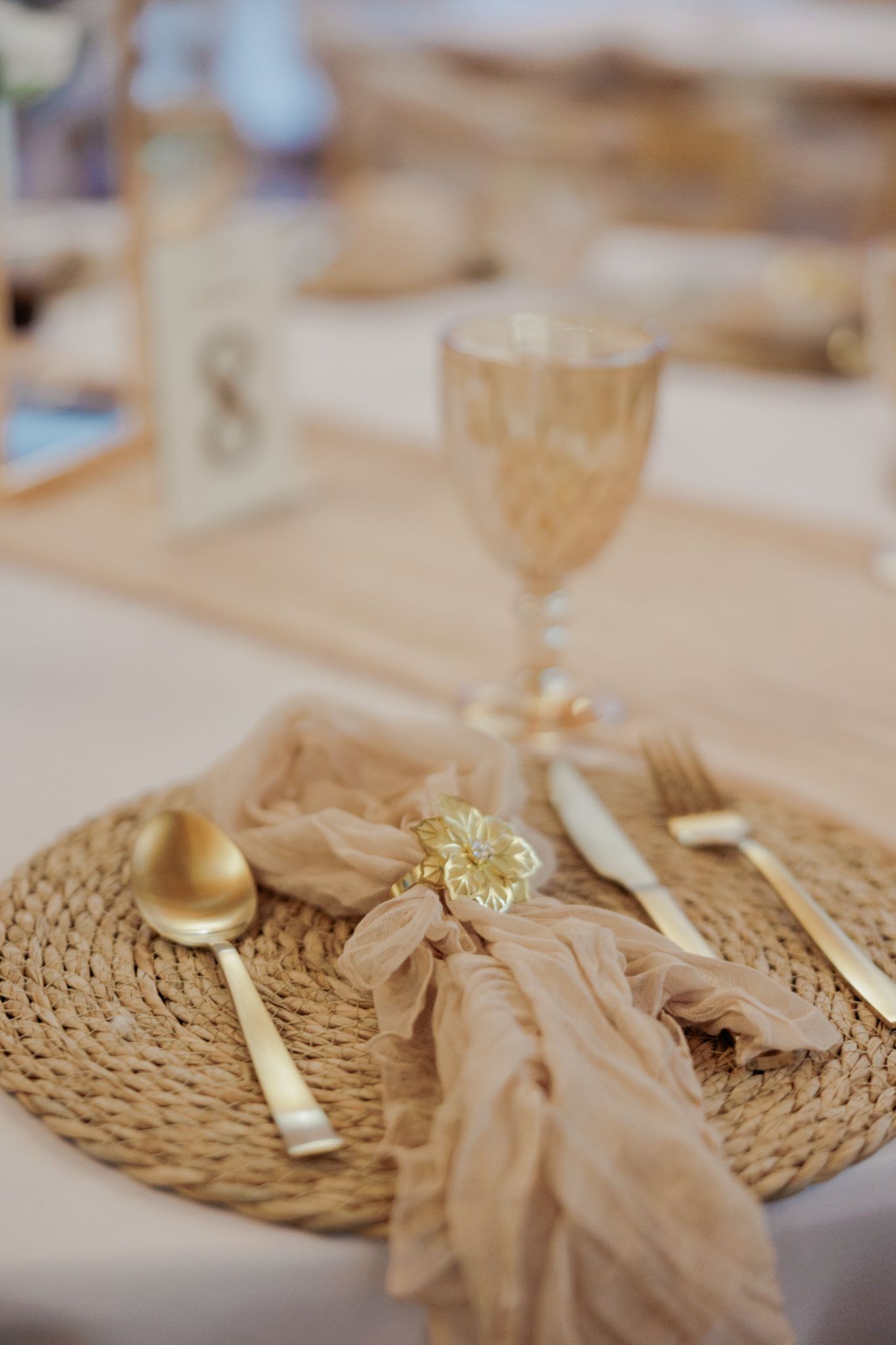 Elegant gold place setting with woven charger, golden cutlery, champagne napkin, and amber goblet for a wedding reception.