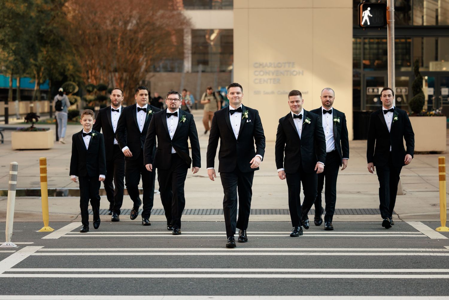 Eight groomsmen in black tuxedos and bow ties walking confidently across a crosswalk in an urban setting.