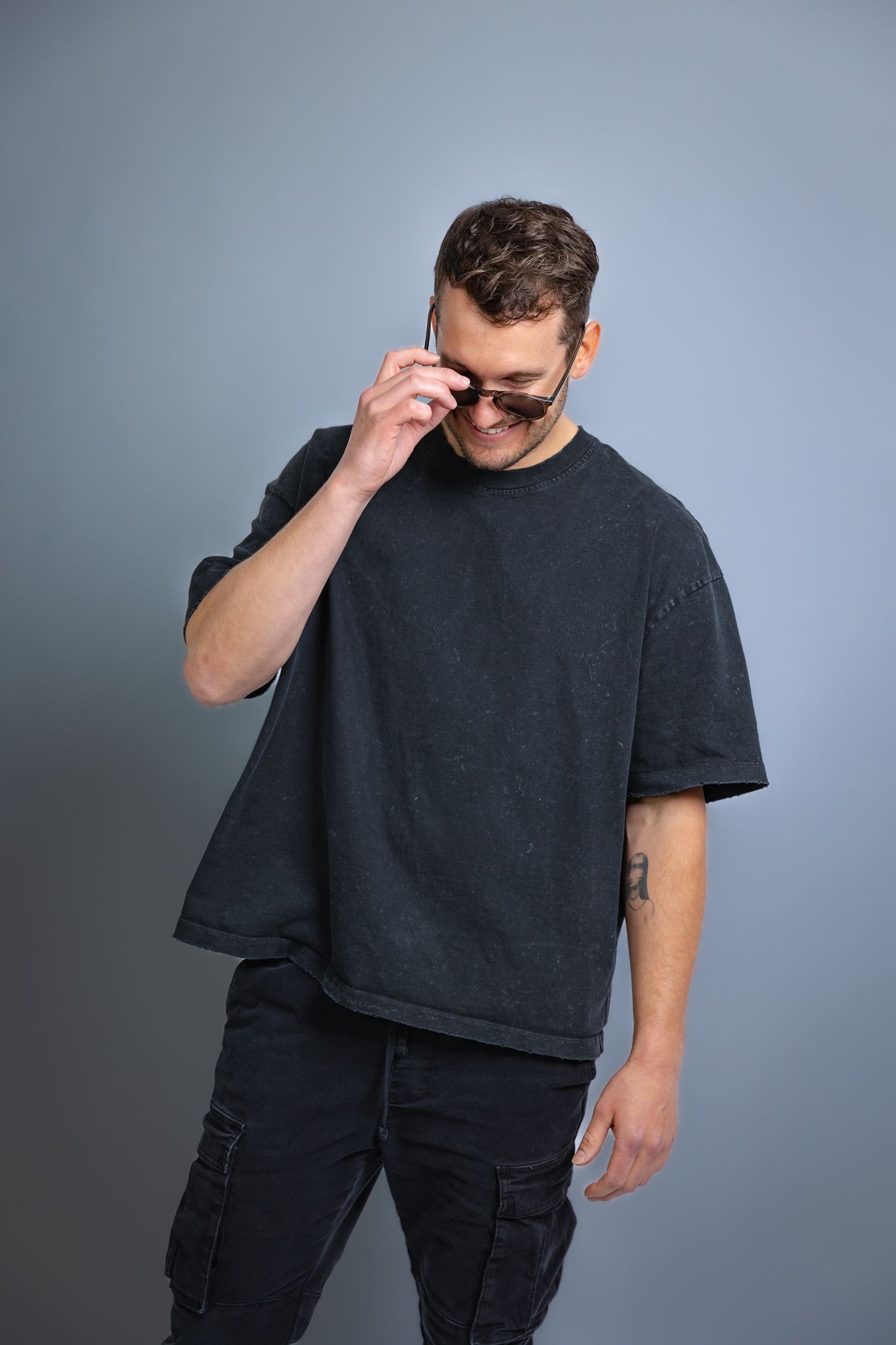 Man in oversized black t-shirt and cargo pants adjusting sunglasses, smiling against gray background.