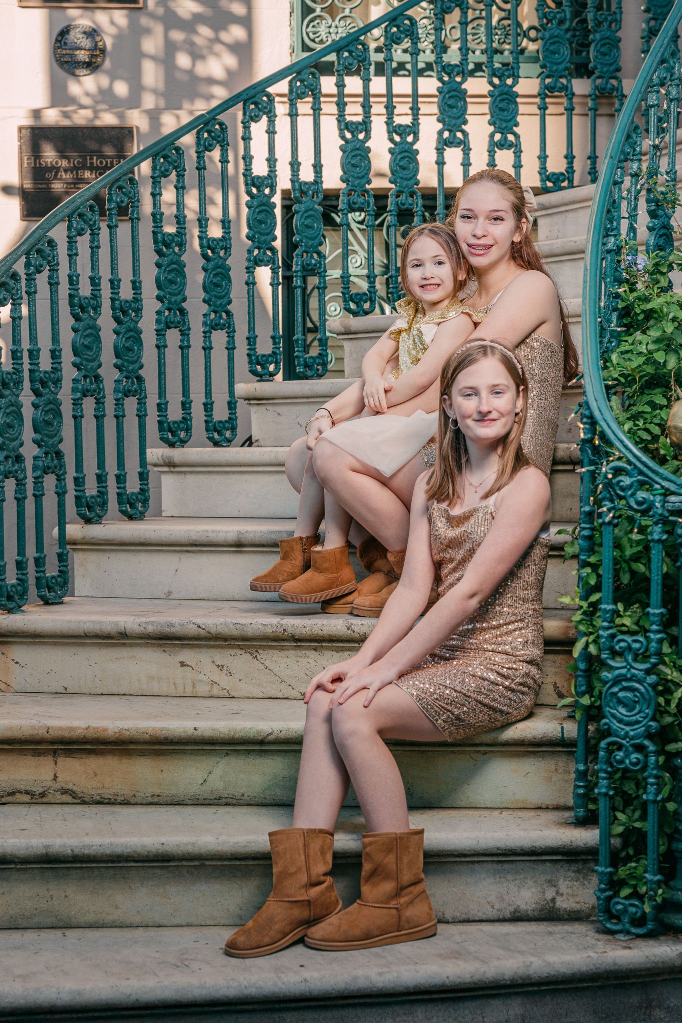 Three girls in gold sequin dresses and brown boots pose on ornate teal iron staircase steps.