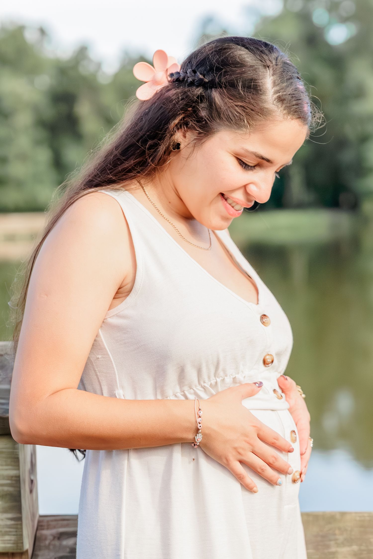 Smiling pregnant woman in white dress cradling her baby bump outdoors near a serene lake on a sunny day.