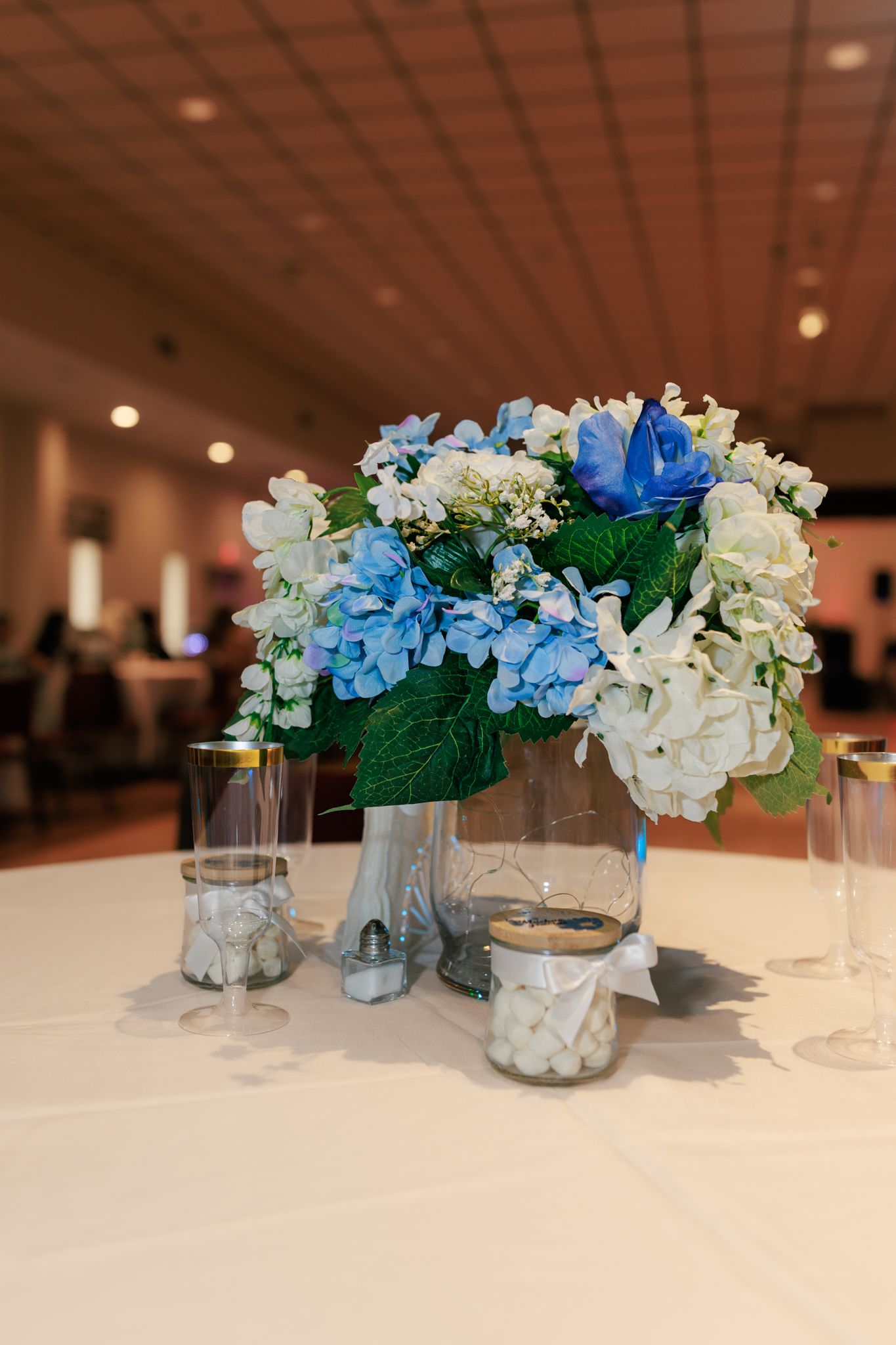 Blue and white hydrangea floral centerpiece in glass vase on white tablecloth at elegant banquet hall.
