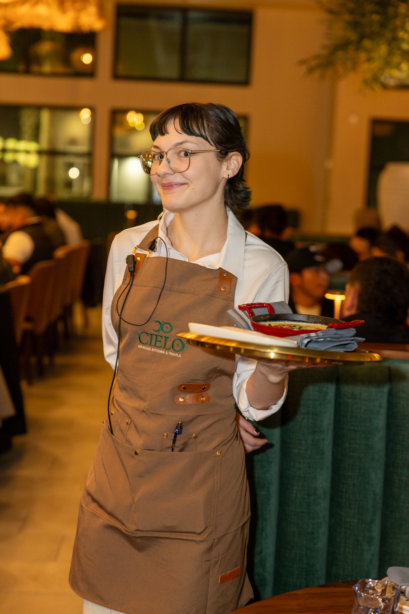 Smiling waitress in brown Cielo restaurant apron carrying a tray in a warmly lit dining room.