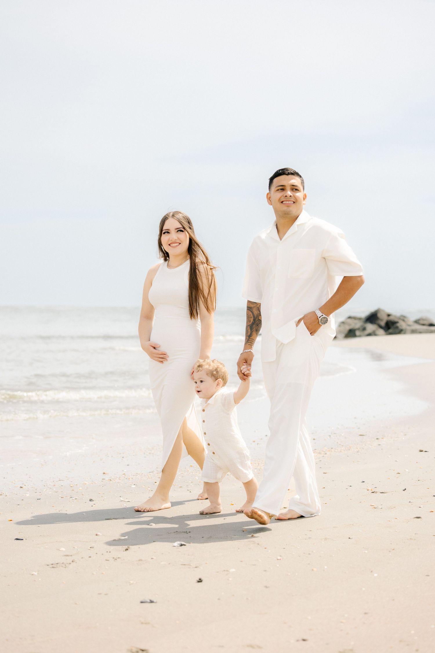 Pregnant family in white outfits walking on beach, mother holding baby toddler hand with father beside them.