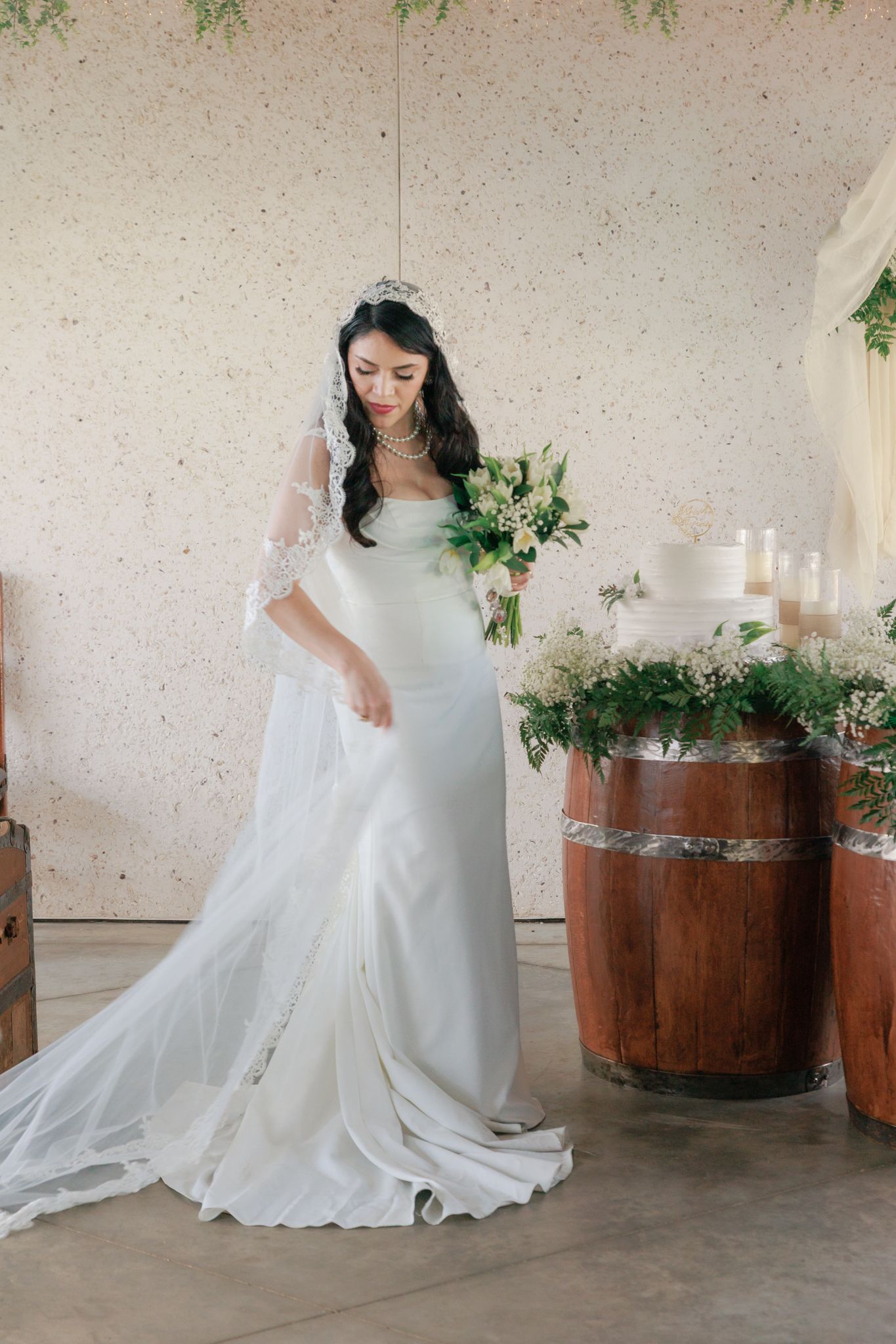 Bride in white gown with lace veil and tiara holds bouquet near rustic wooden barrels at elegant wedding venue.