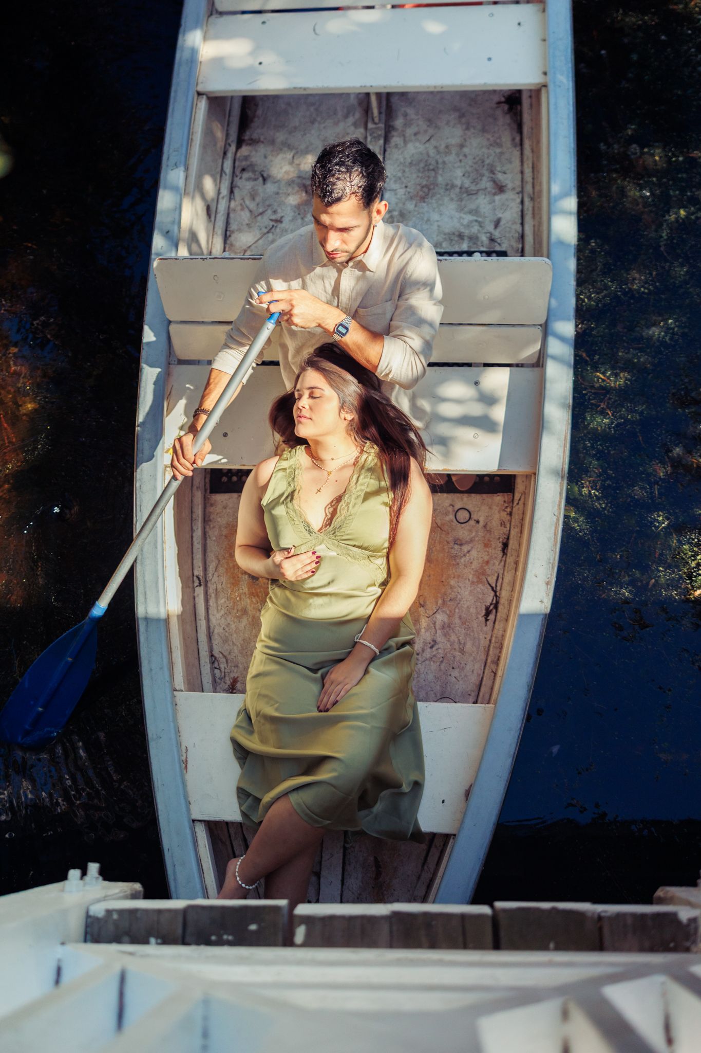 Romantic couple relaxing in a wooden rowboat on calm water, woman in green dress leaning back while man holds oar.