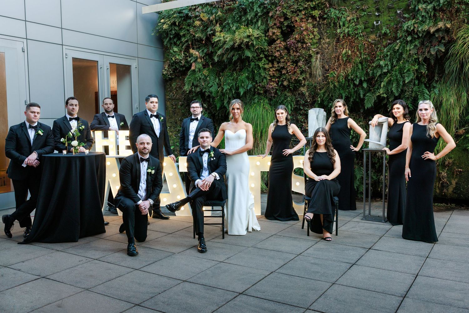 Wedding party in black attire poses outdoors against a lush green wall with marquee letters at elegant venue.