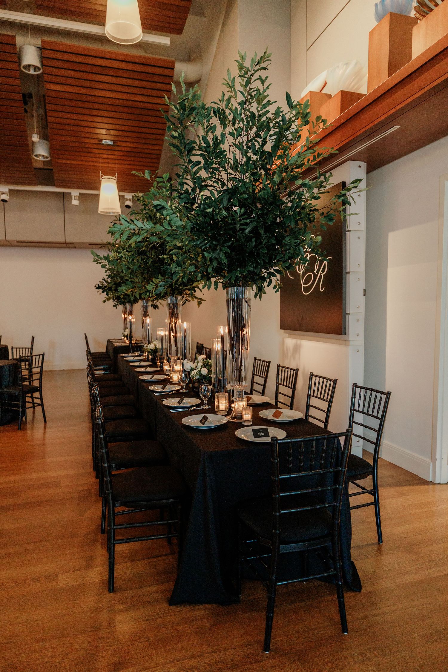 Elegant wedding reception with long black table, tall greenery centerpiece, candles, and black Chiavari chairs in modern venue.