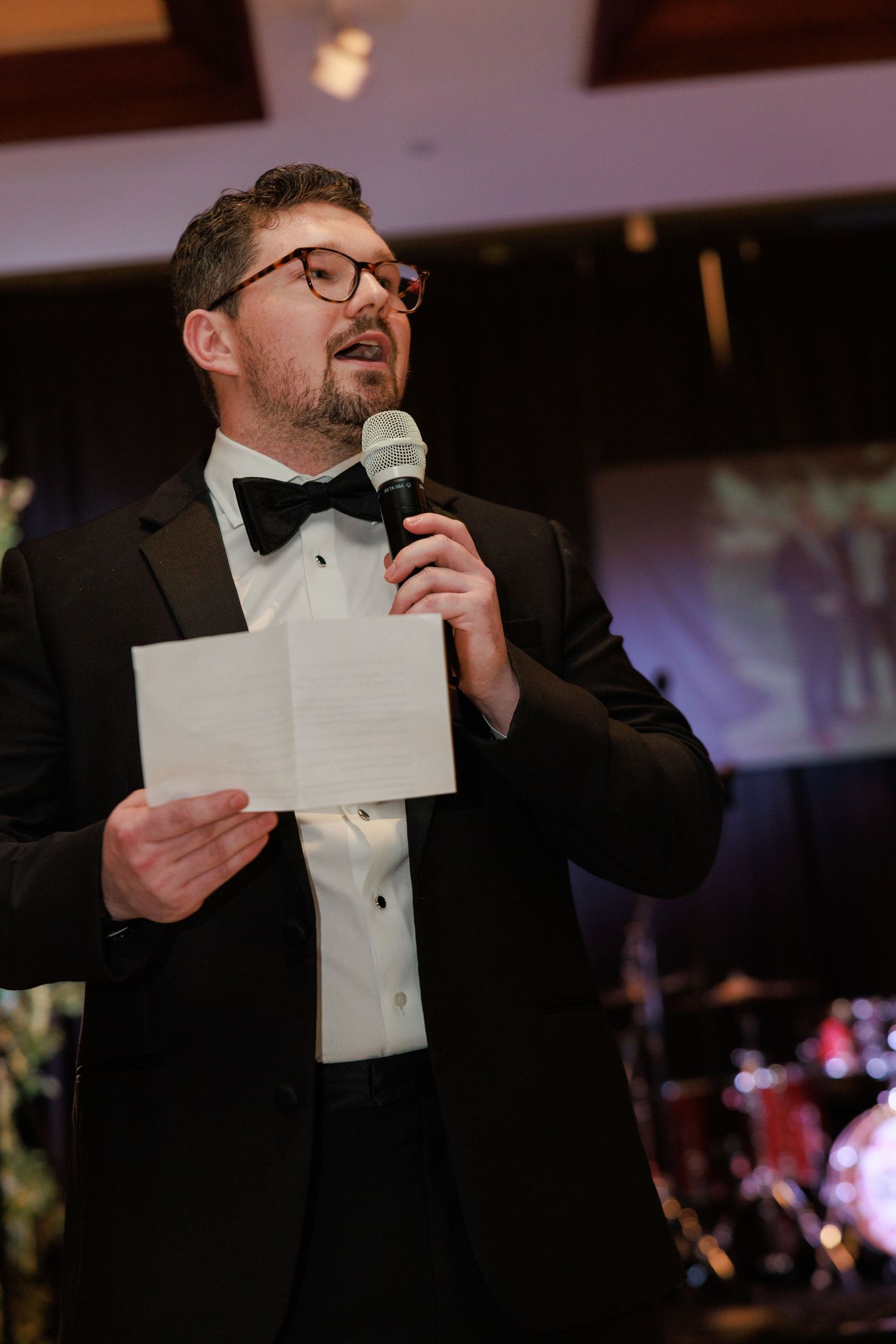 Man in tuxedo and bow tie delivering speech at formal event, holding microphone and notes.