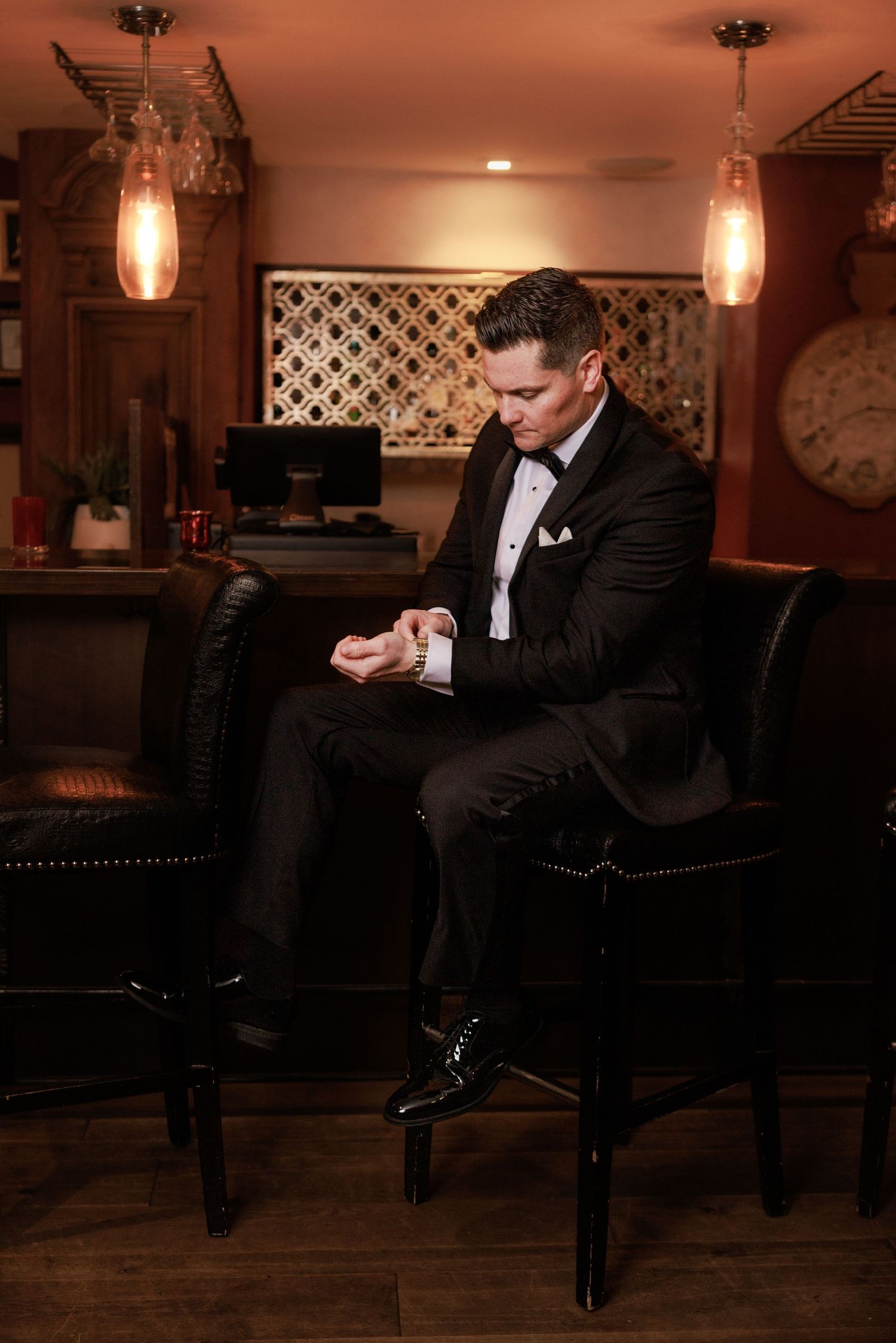 A well-dressed groom in a black tuxedo adjusts his cufflinks while seated in a leather chair in a warmly lit bar.