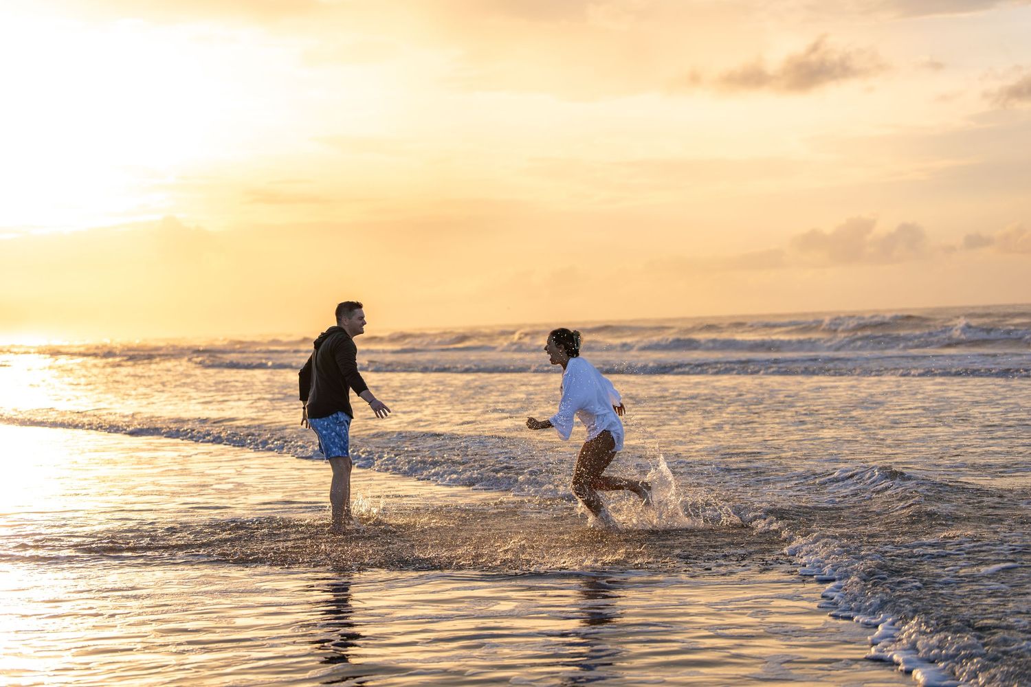 Playful couple splashing in ocean waves during golden hour sunset.