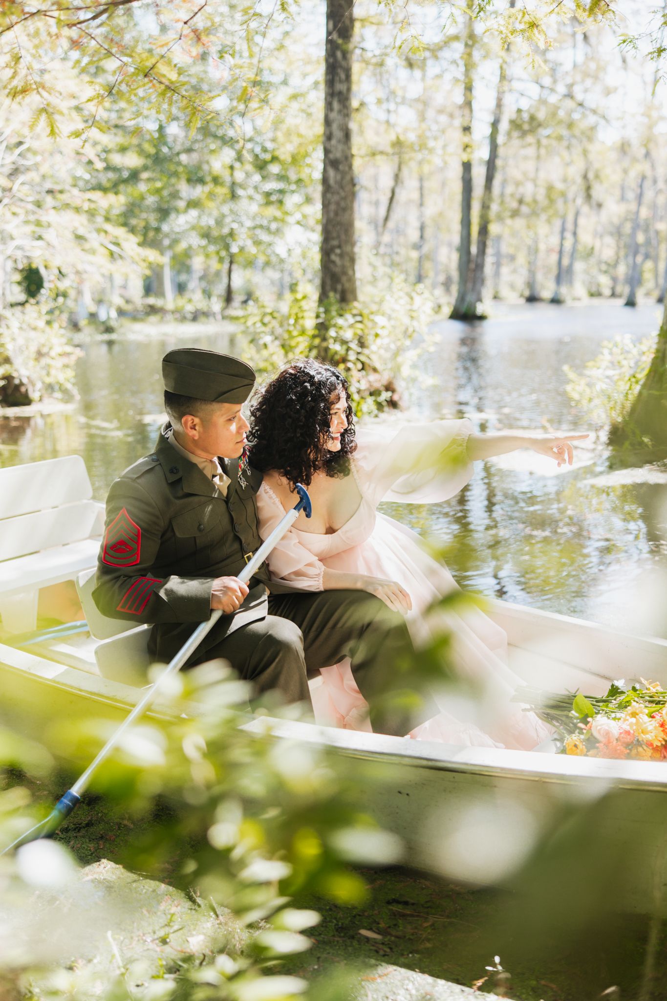 A couple in a white rowboat on a serene pond, man in military uniform, woman in pink dress, surrounded by lush trees.