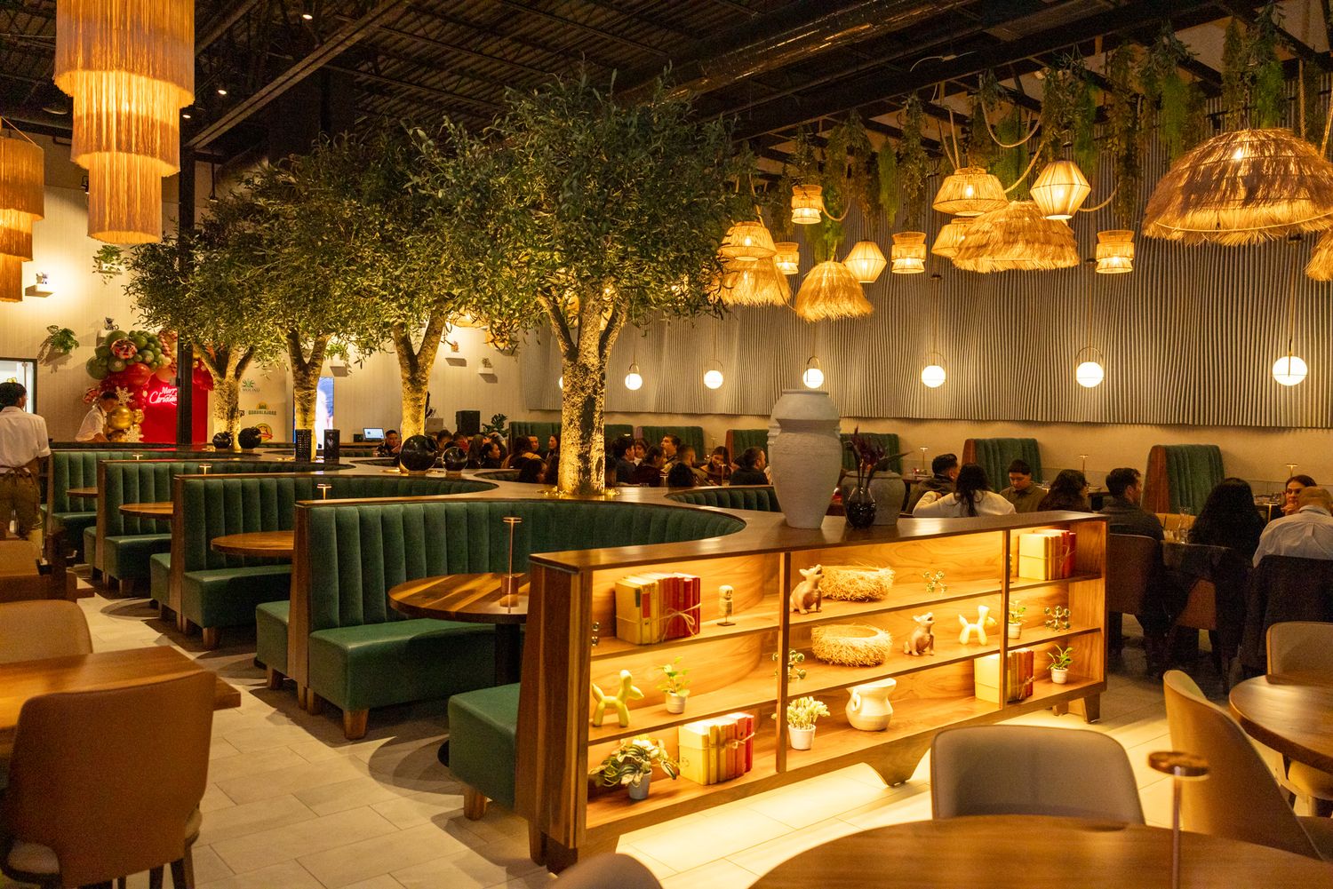 Elegant restaurant interior with green velvet booths, wooden shelving, and warm pendant lights amid lush indoor trees.